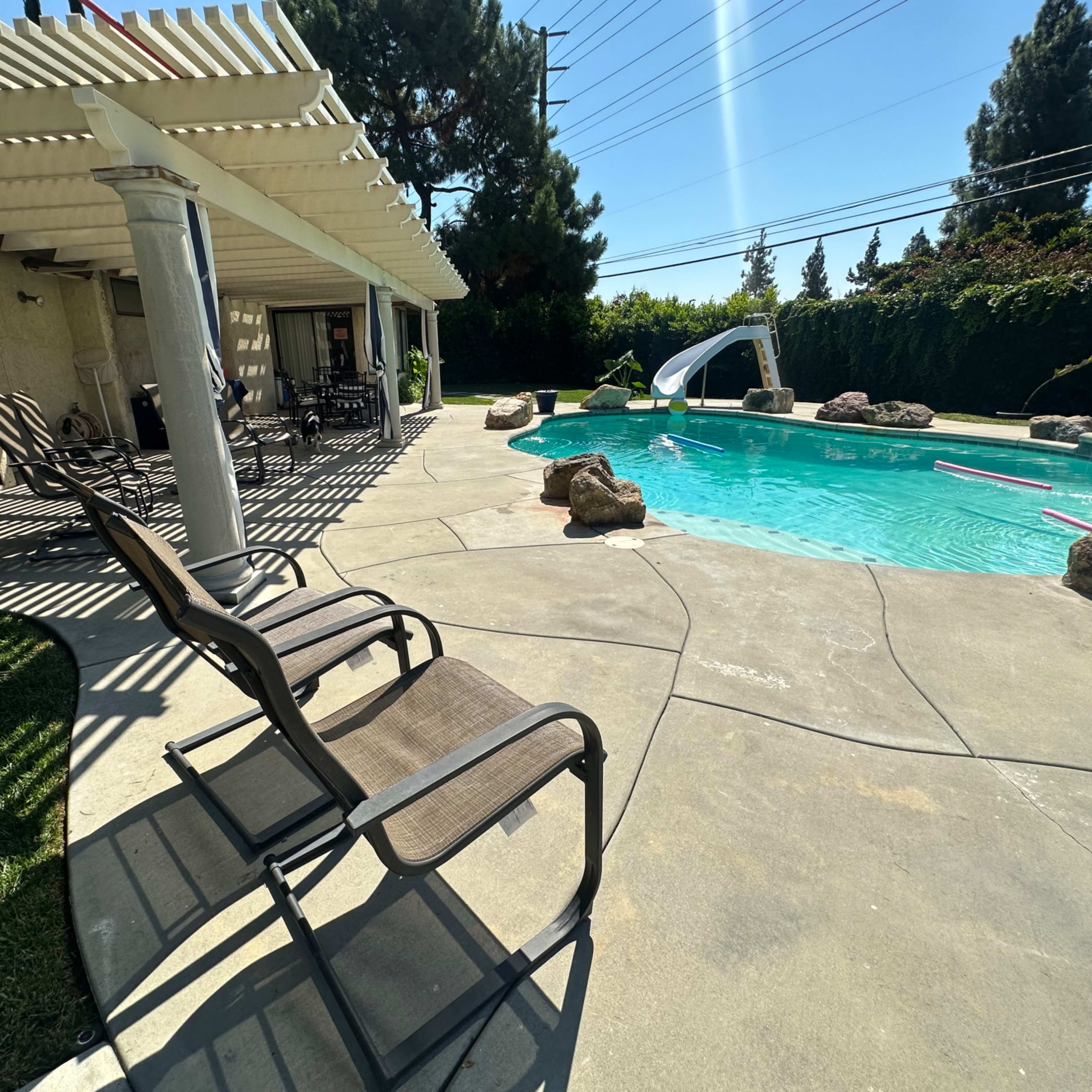 A backyard pool area with a slide, lounge chairs, and a patio surrounded by greenery.