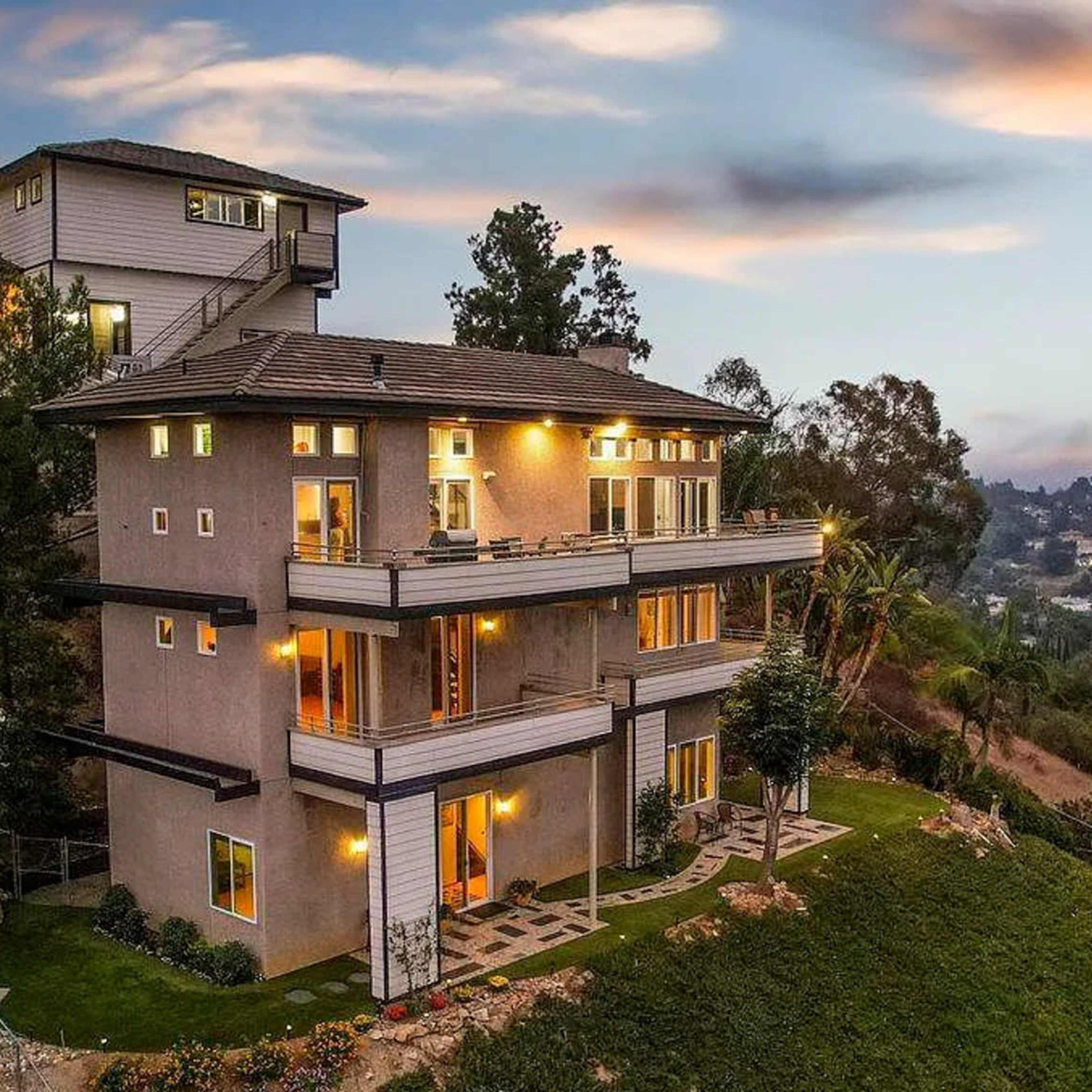 A modern multi-level house perched on a hillside, surrounded by trees and gardens, with lights glowing in the windows as dusk approaches.
