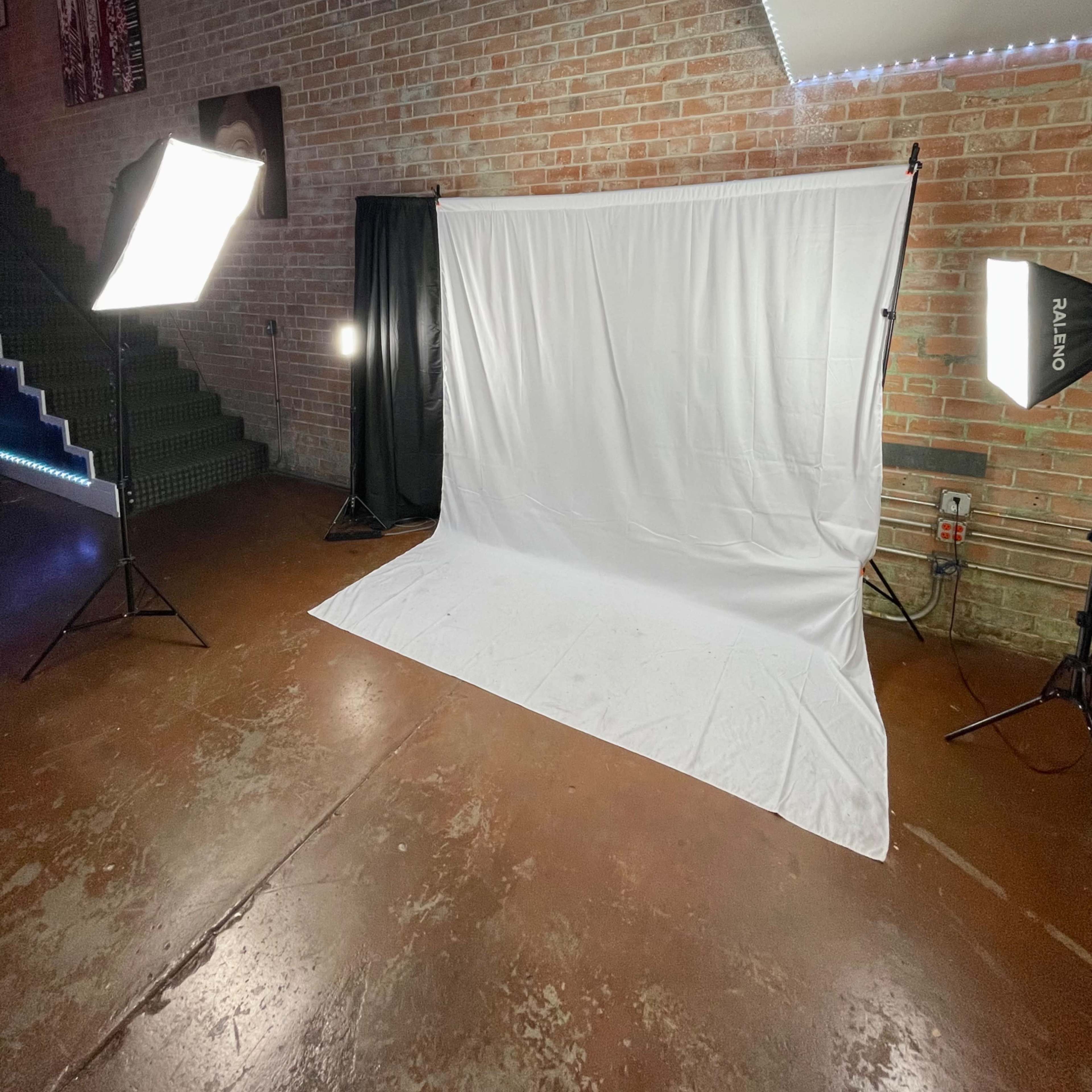 A spacious room features a white backdrop with two bright lights positioned on stands nearby, alongside a staircase in the background.