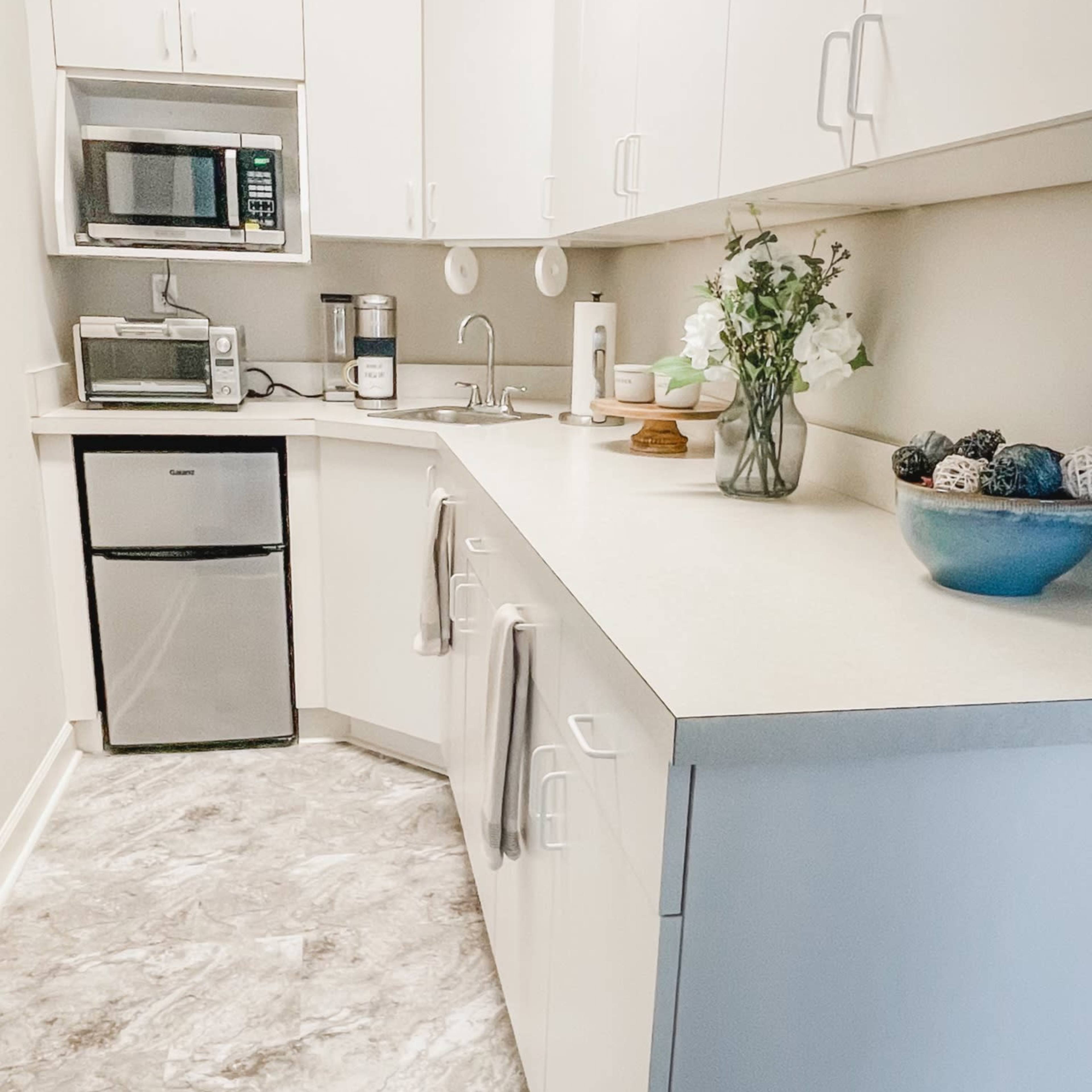 A compact kitchen features white cabinets, a stainless steel microwave, a mini refrigerator, and a countertop adorned with a vase of flowers and decorative items.