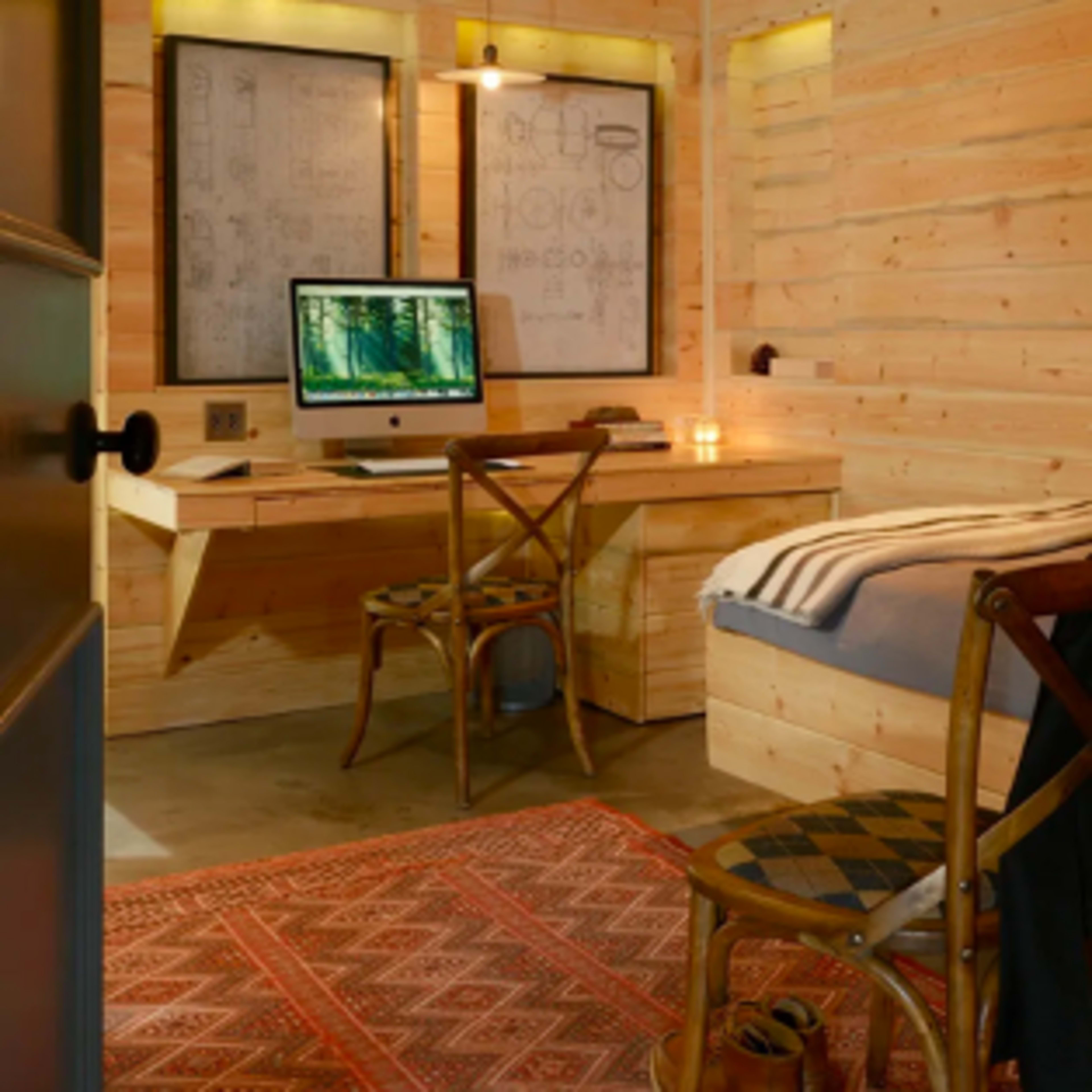 A rustic wooden office space with a desk, computer, and a bed, all illuminated by warm lighting.