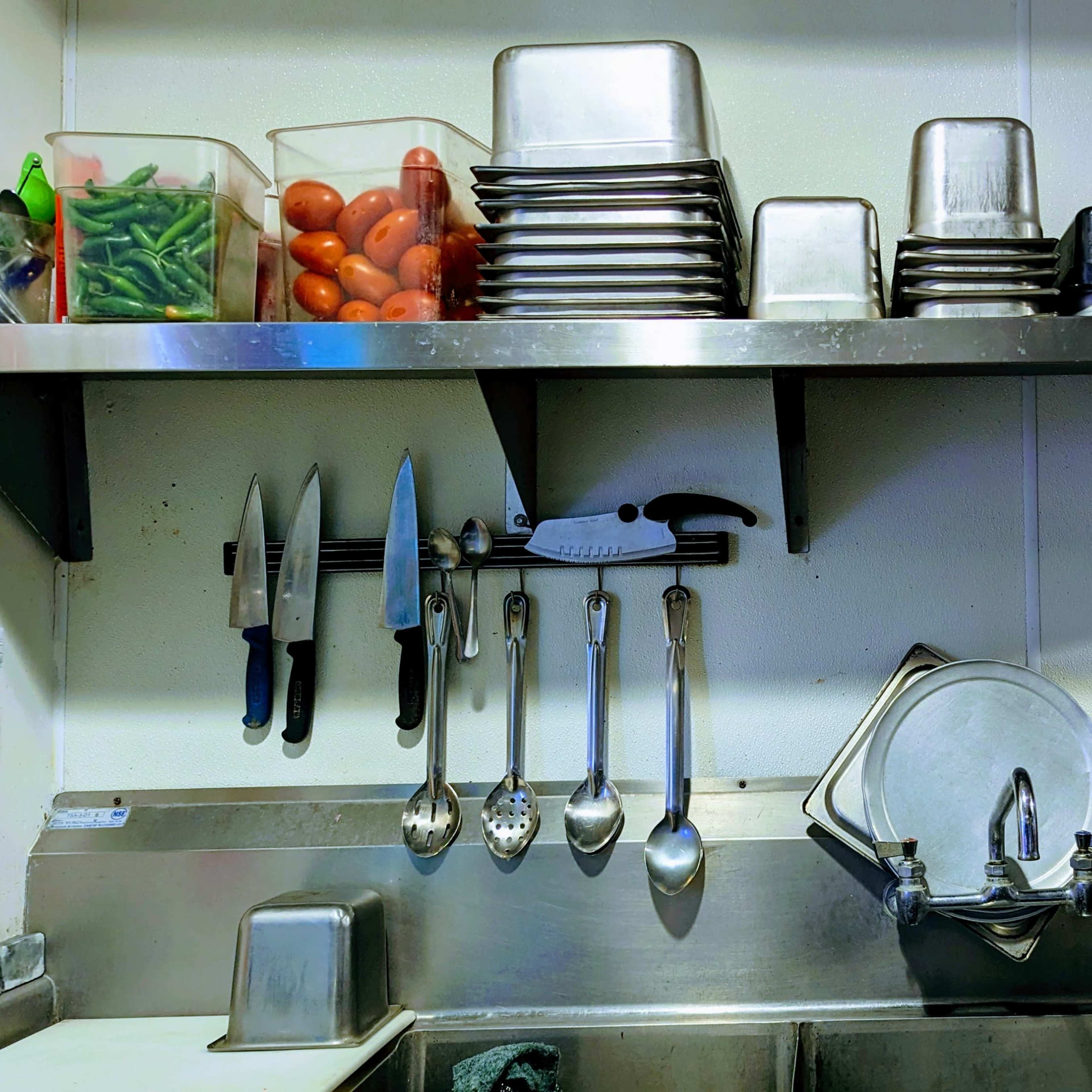 A kitchen workspace with metal shelving holding containers of vegetables, plates, and utensils, along with hanging knives and various tools near a stainless steel sink.