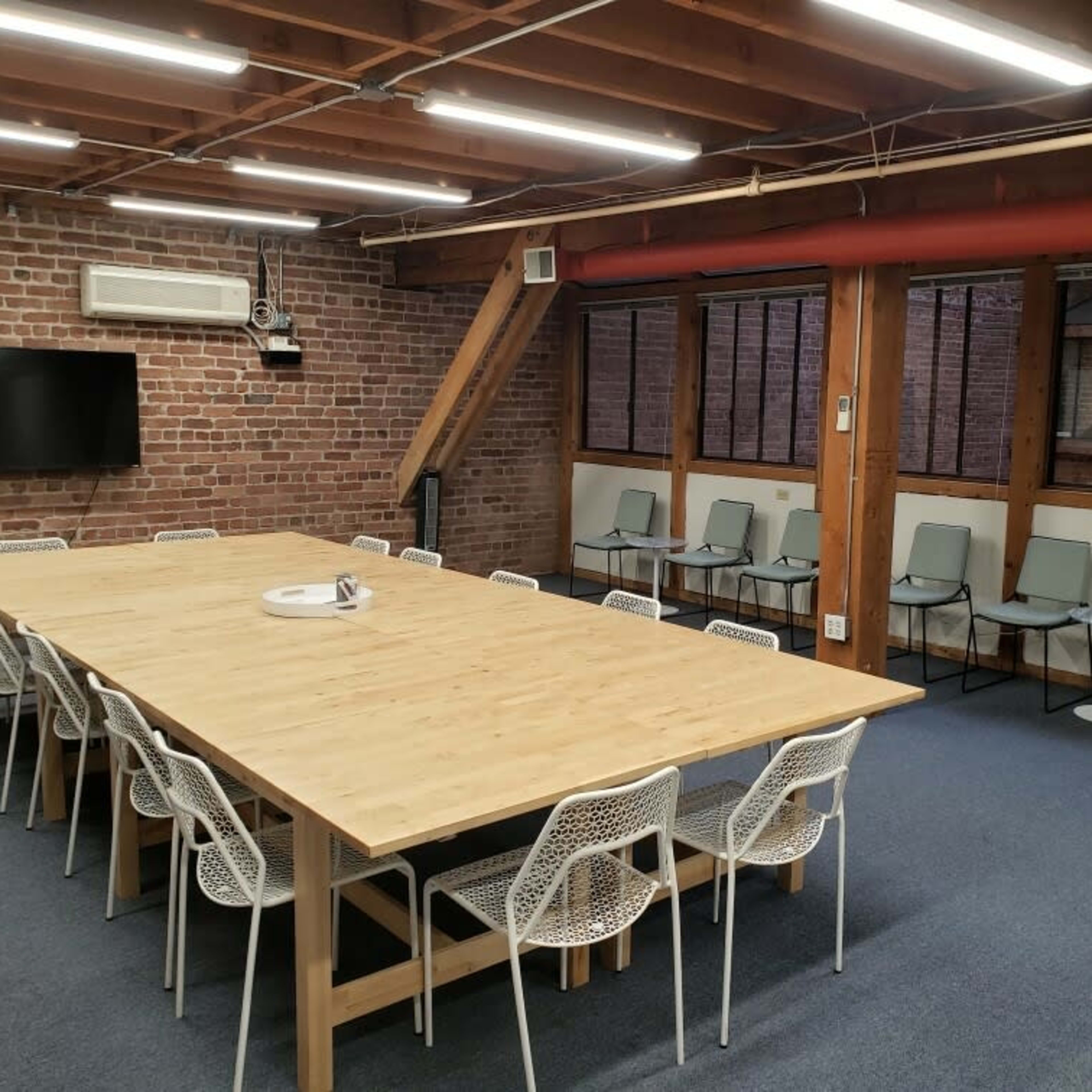 A large wooden table surrounded by white chairs occupies the center of a meeting room with exposed brick walls and large windows.