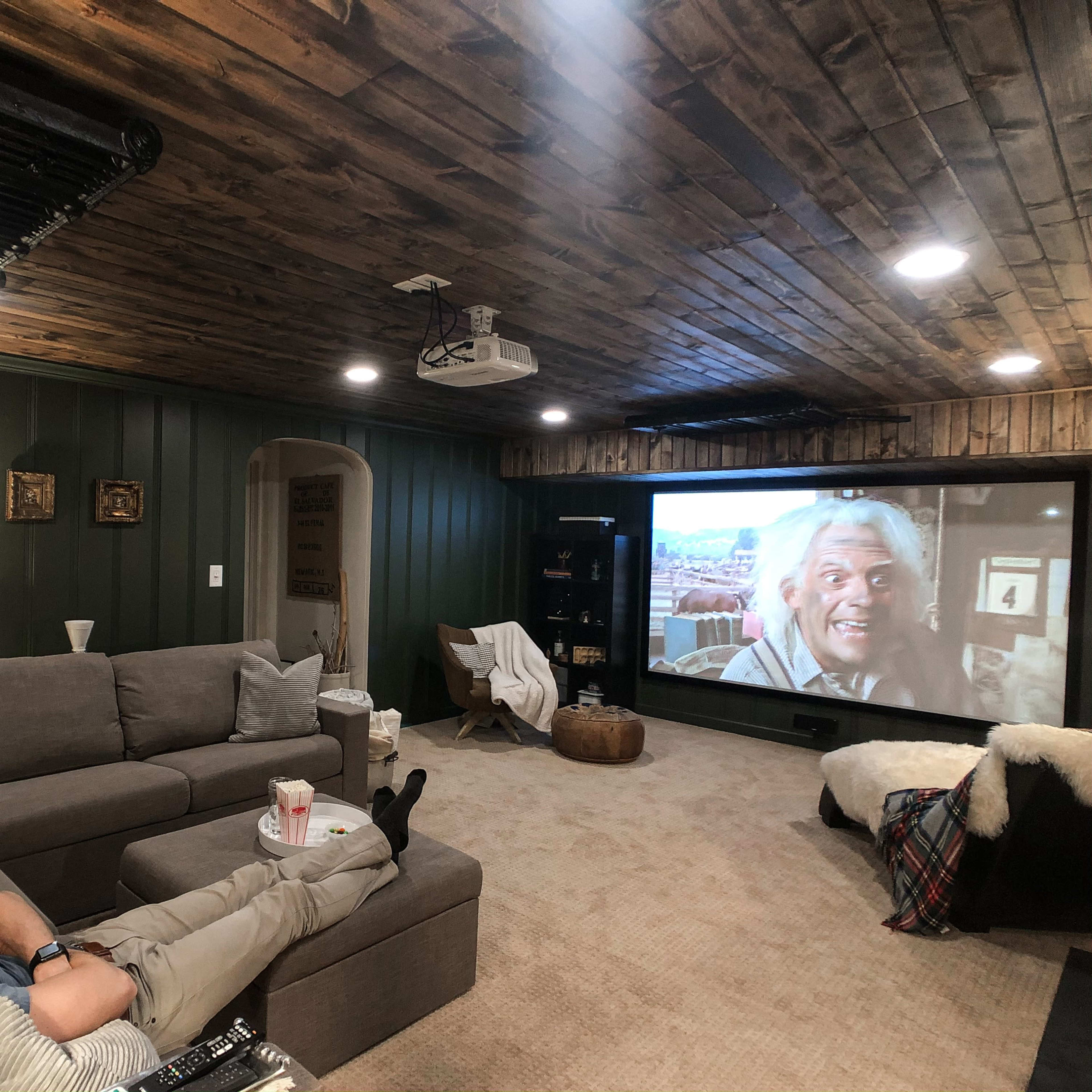 A person reclines on a gray sectional couch in a cozy basement watching a movie on a large projection screen.
