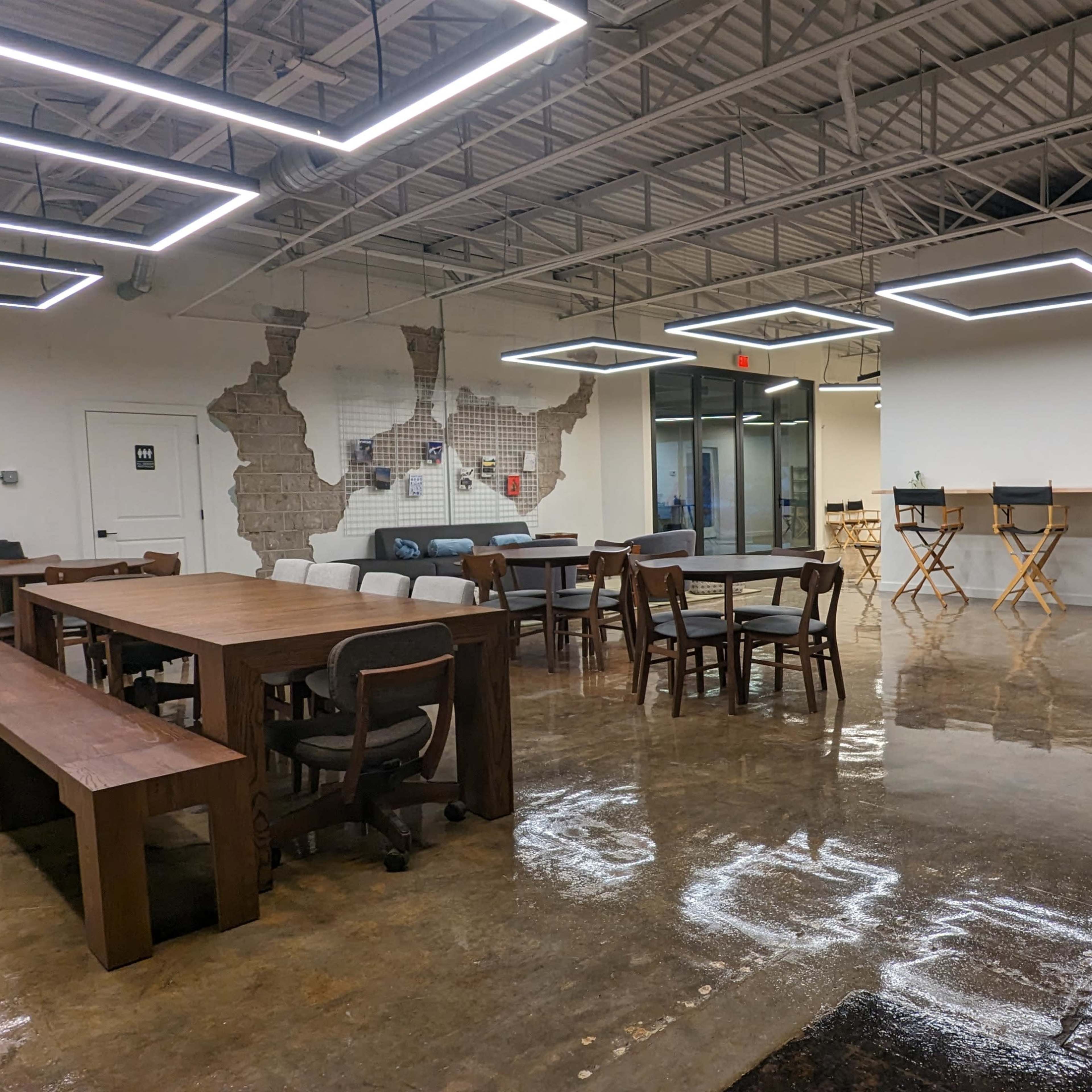A modern, open workspace featuring a mix of wooden furniture, including dining tables and chairs, with polished concrete floors and stylish lighting fixtures.