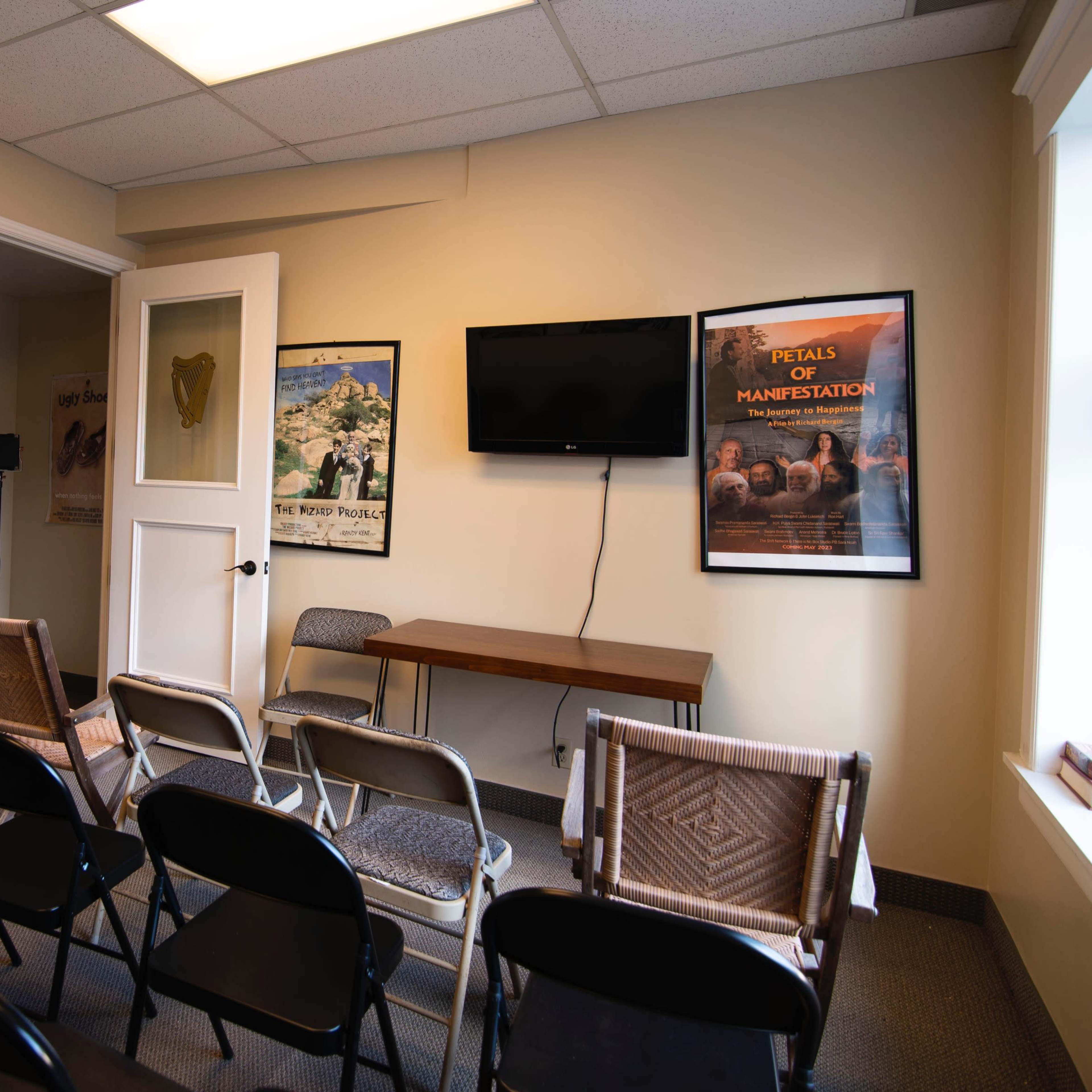 A small meeting room with rows of folding chairs facing a wall-mounted television and a table against the opposite wall, adorned with posters.