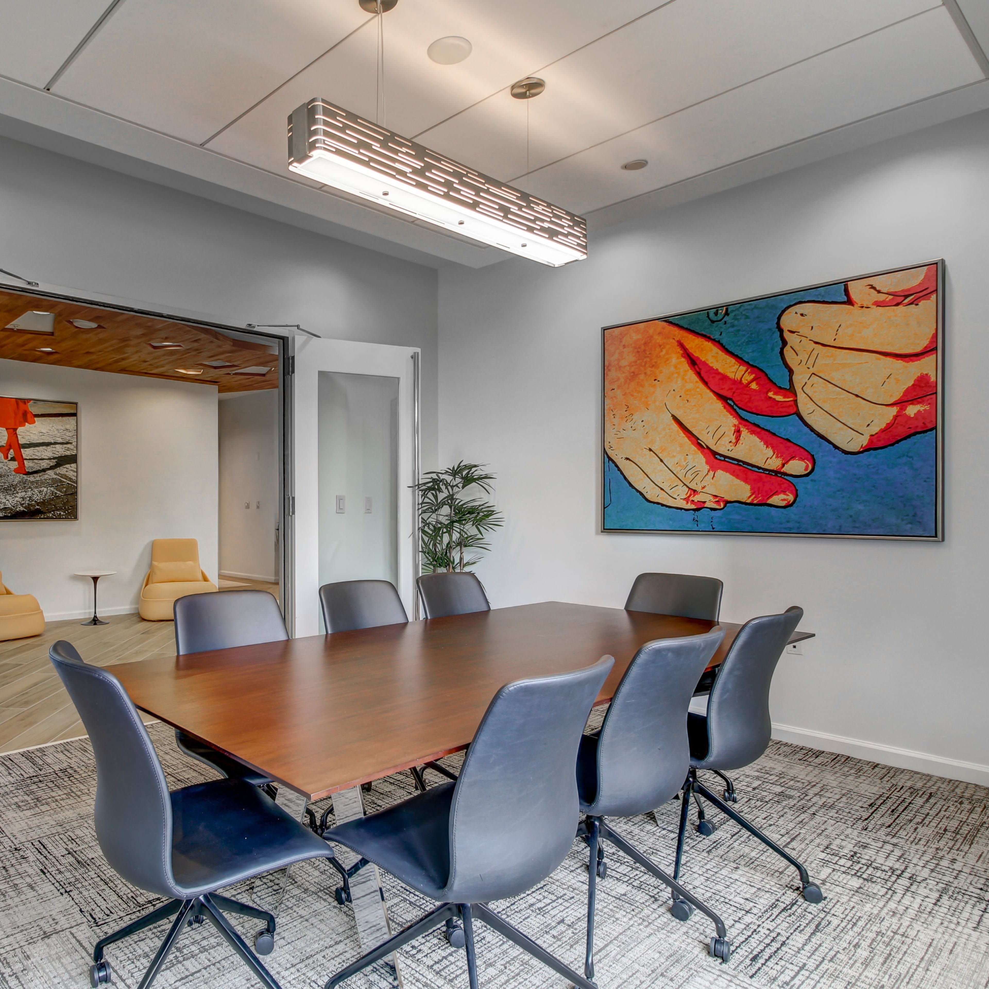 A conference room features a large wooden table surrounded by black chairs, with a contemporary artwork on the wall and a view into a lounge area through open double doors.