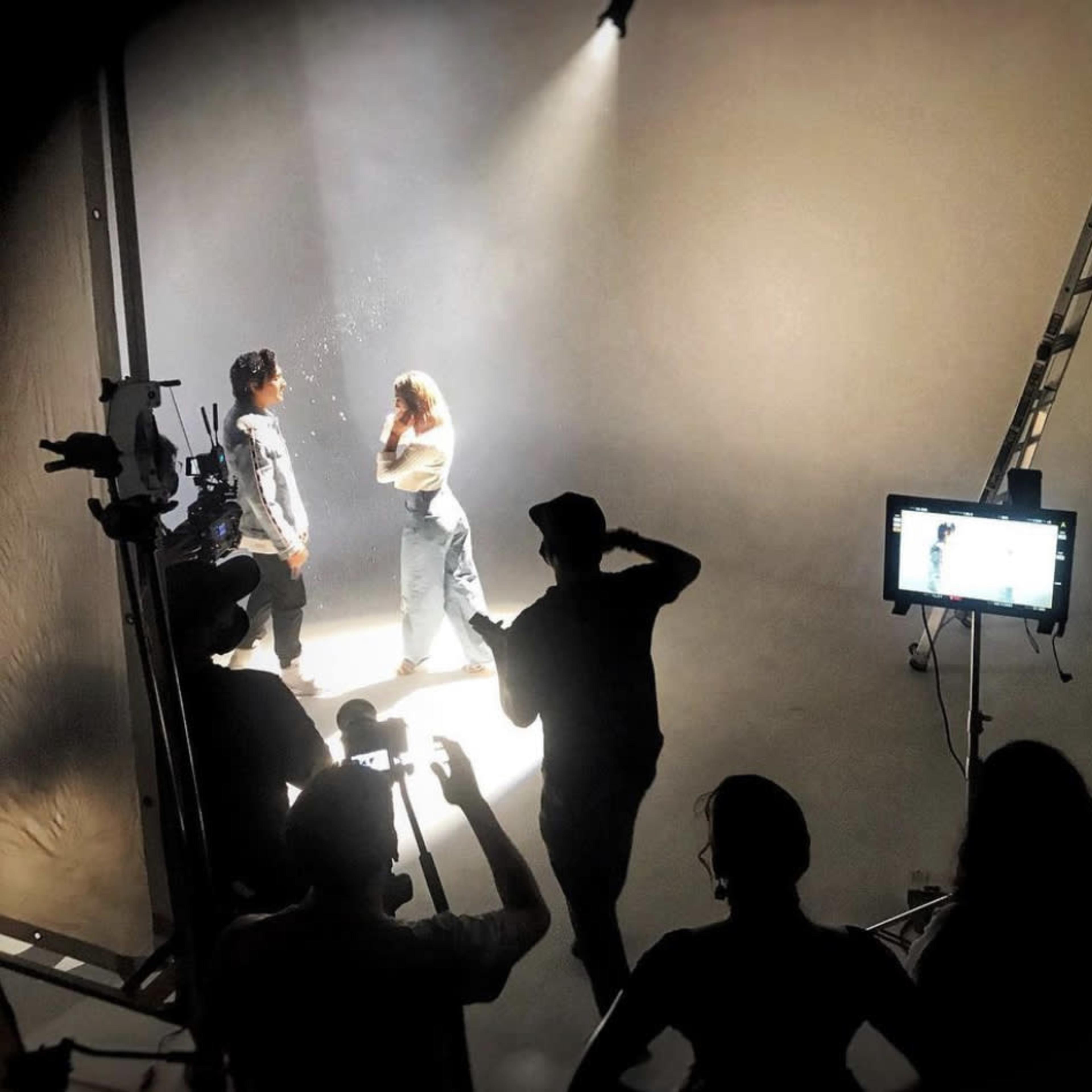 A group of people observes the filming of a scene featuring two models illuminated by soft lighting in a studio setting.