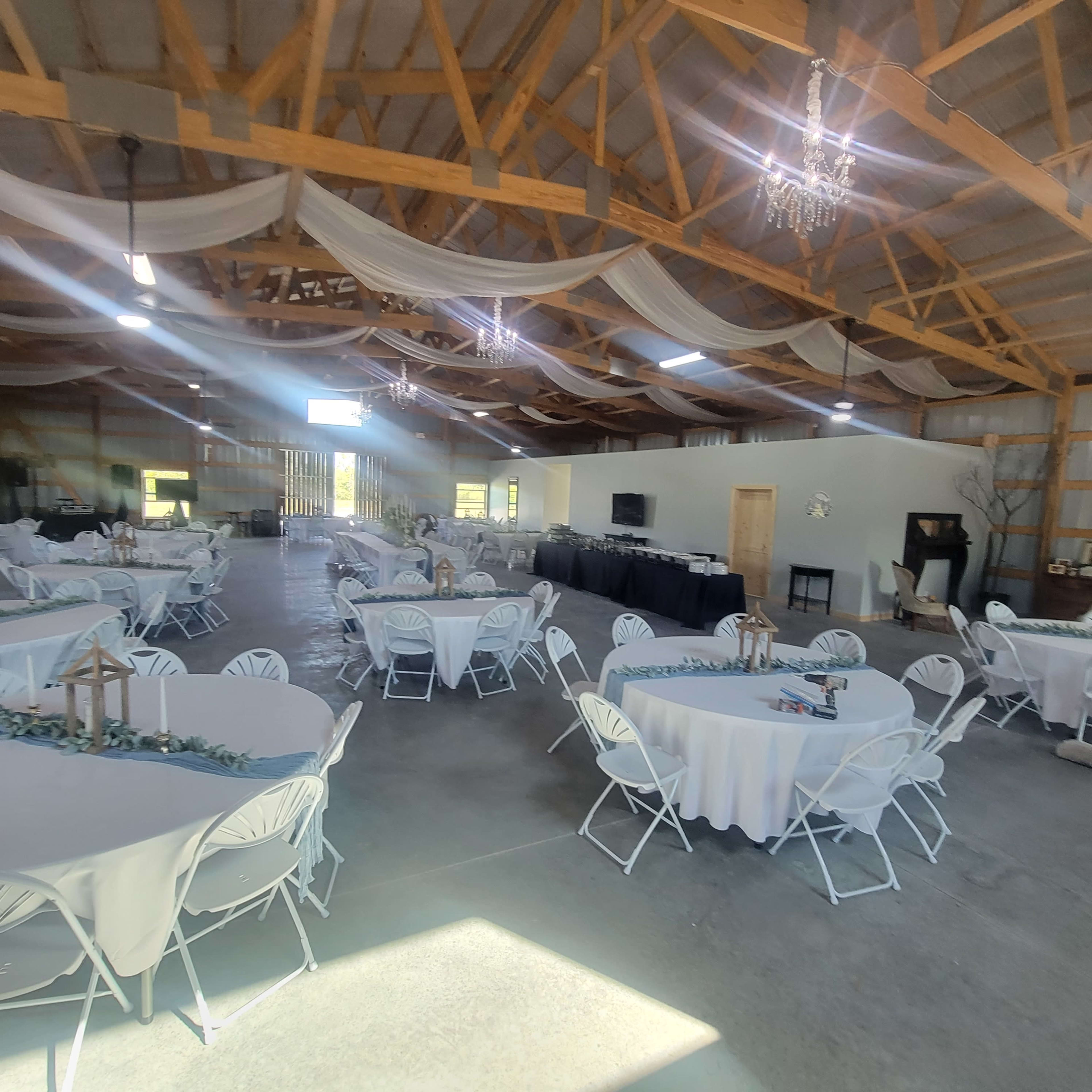 A large, open event space with round tables covered in white tablecloths, arranged neatly under a wooden ceiling with chandeliers.