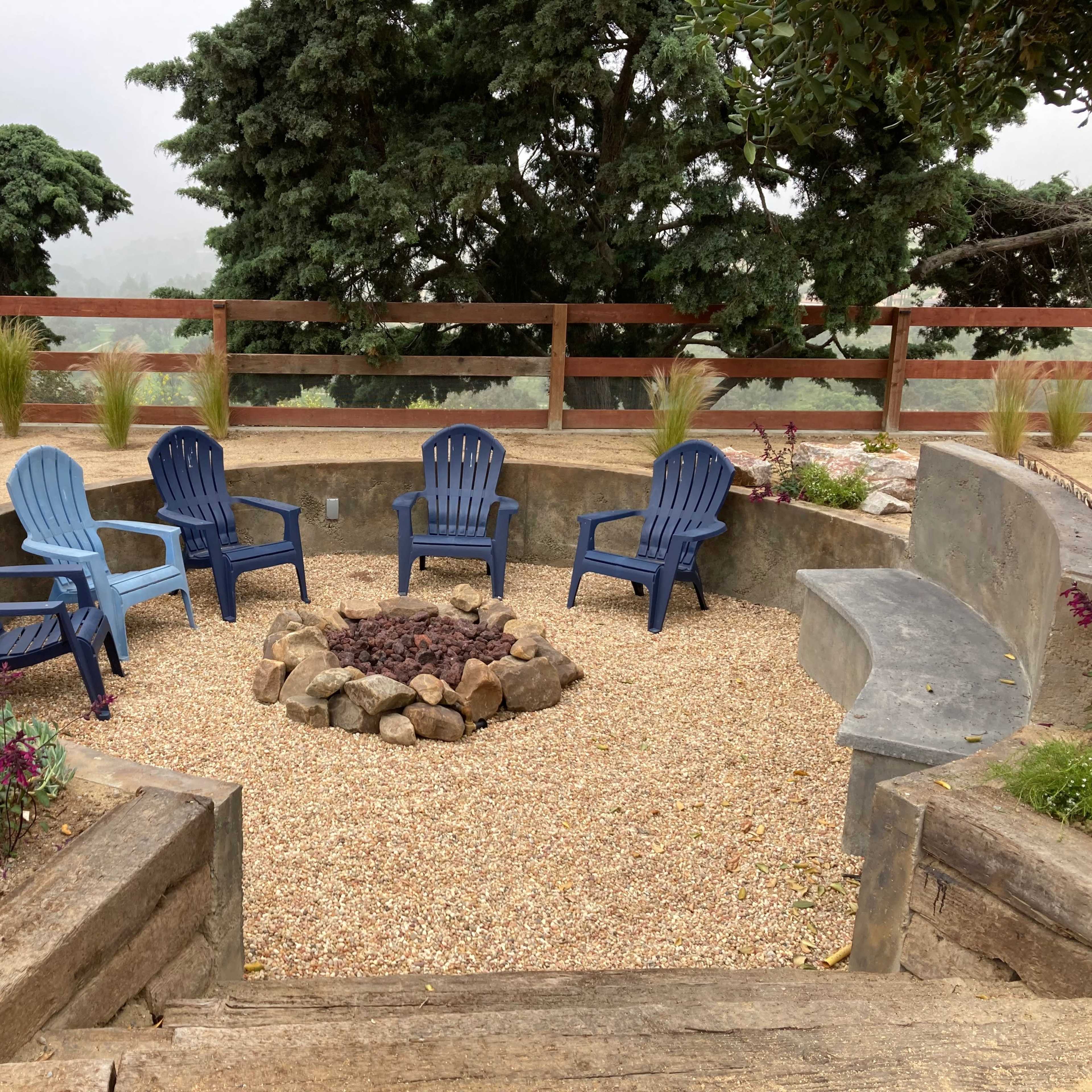 A circular seating area surrounded by gravel features five blue Adirondack chairs arranged around a stone fire pit.