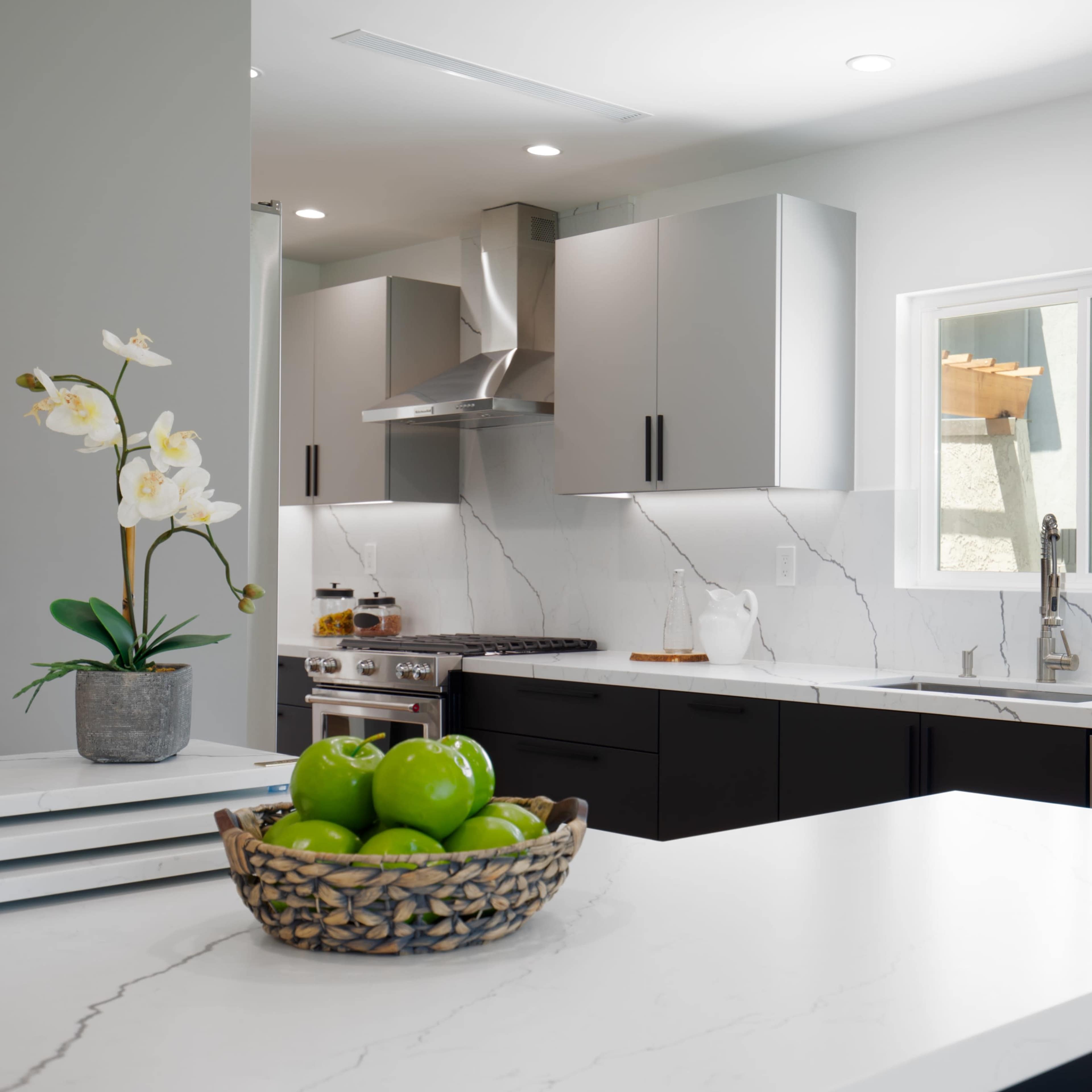 A modern kitchen features sleek gray and black cabinetry, a white marble countertop, a bowl of green apples, and a window overlooking an outdoor area.