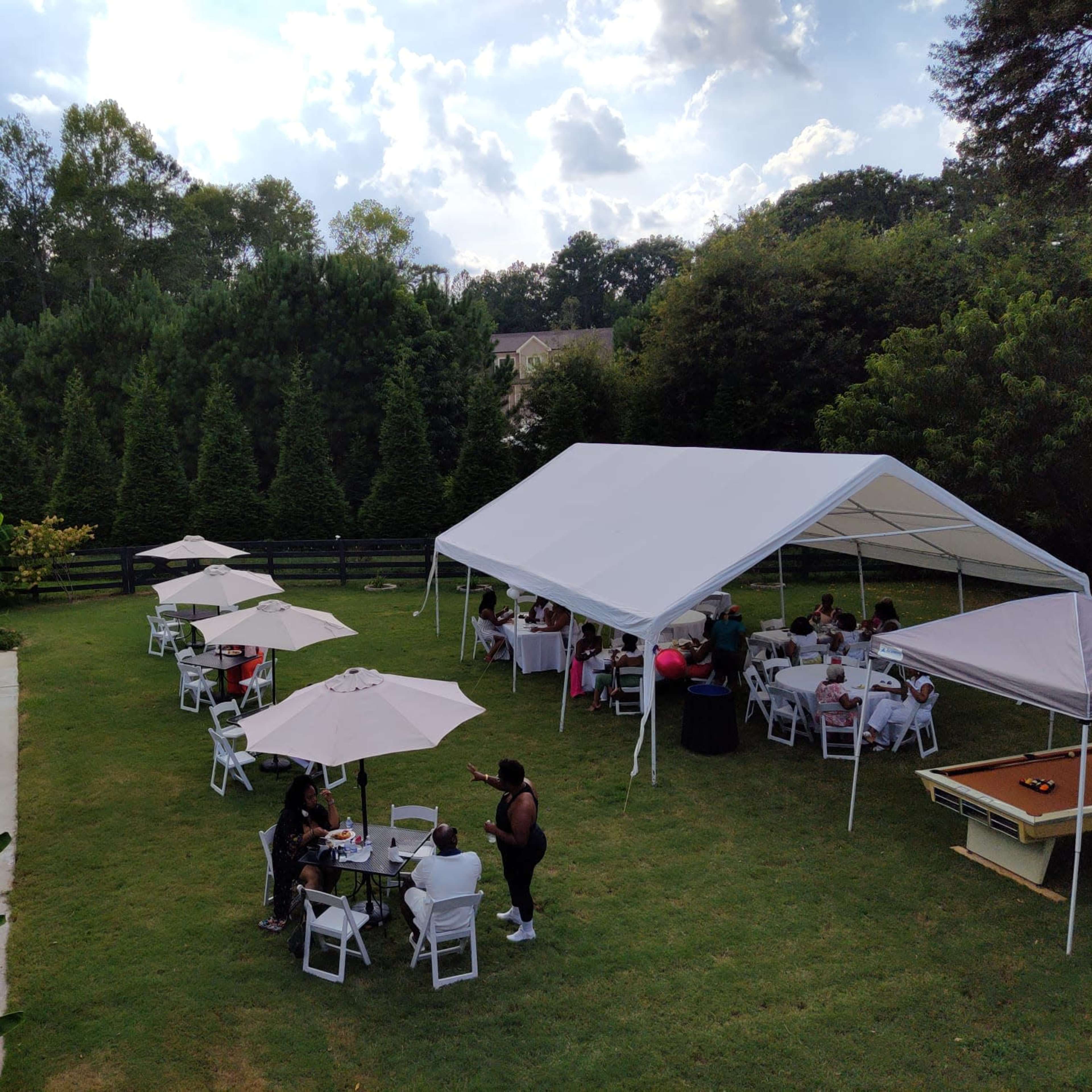 A large outdoor gathering takes place under a tent near a swimming pool, with guests seated around tables and umbrellas in a grassy area.