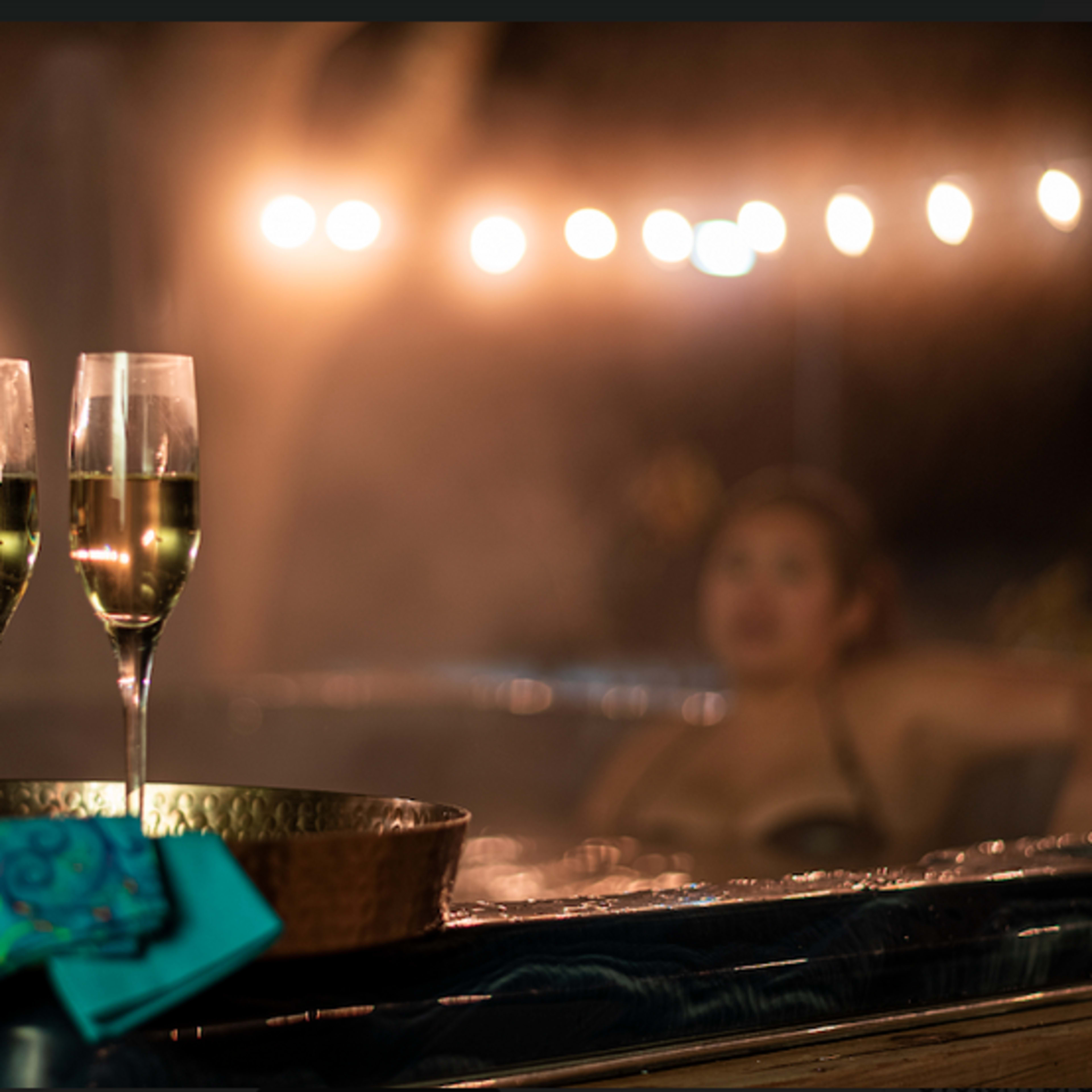 A bottle of sparkling wine and two fluted glasses are set on a tray beside a hot tub, with soft lighting in the background.