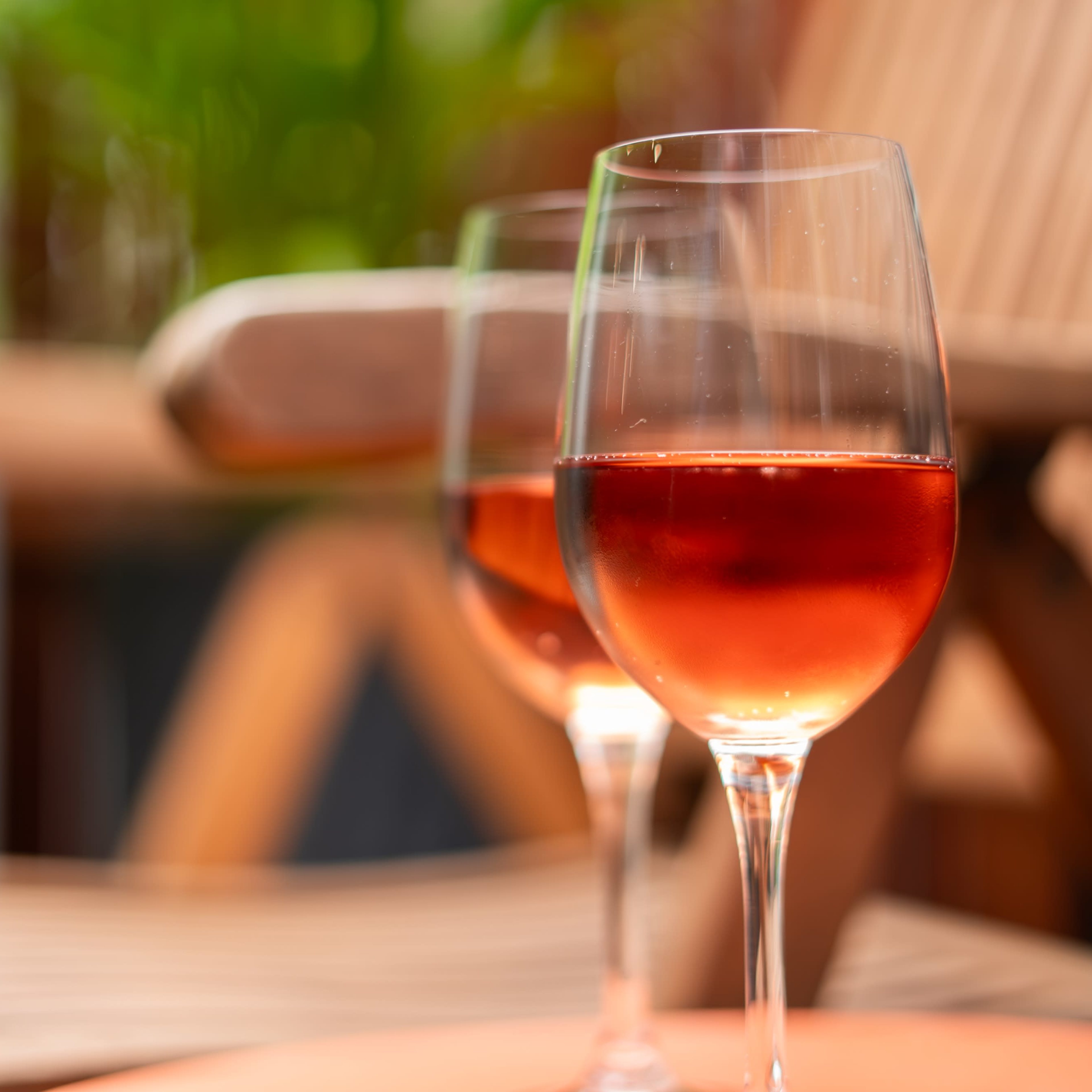 Two glasses of rosé wine sit on a table near a wooden chair in a lush, green setting.