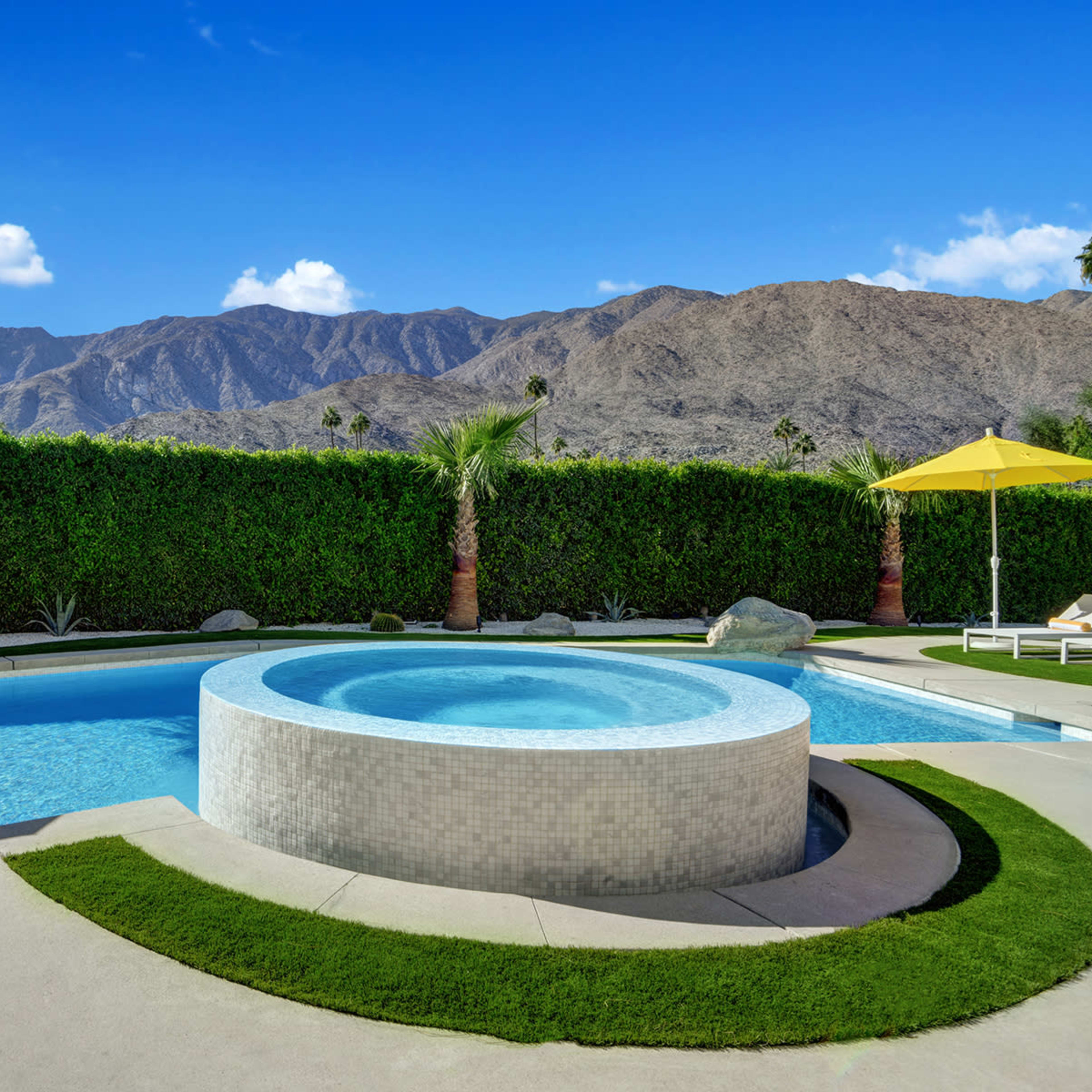 A modern swimming pool with a hot tub, surrounded by green palm trees and a mountain backdrop under a clear blue sky.