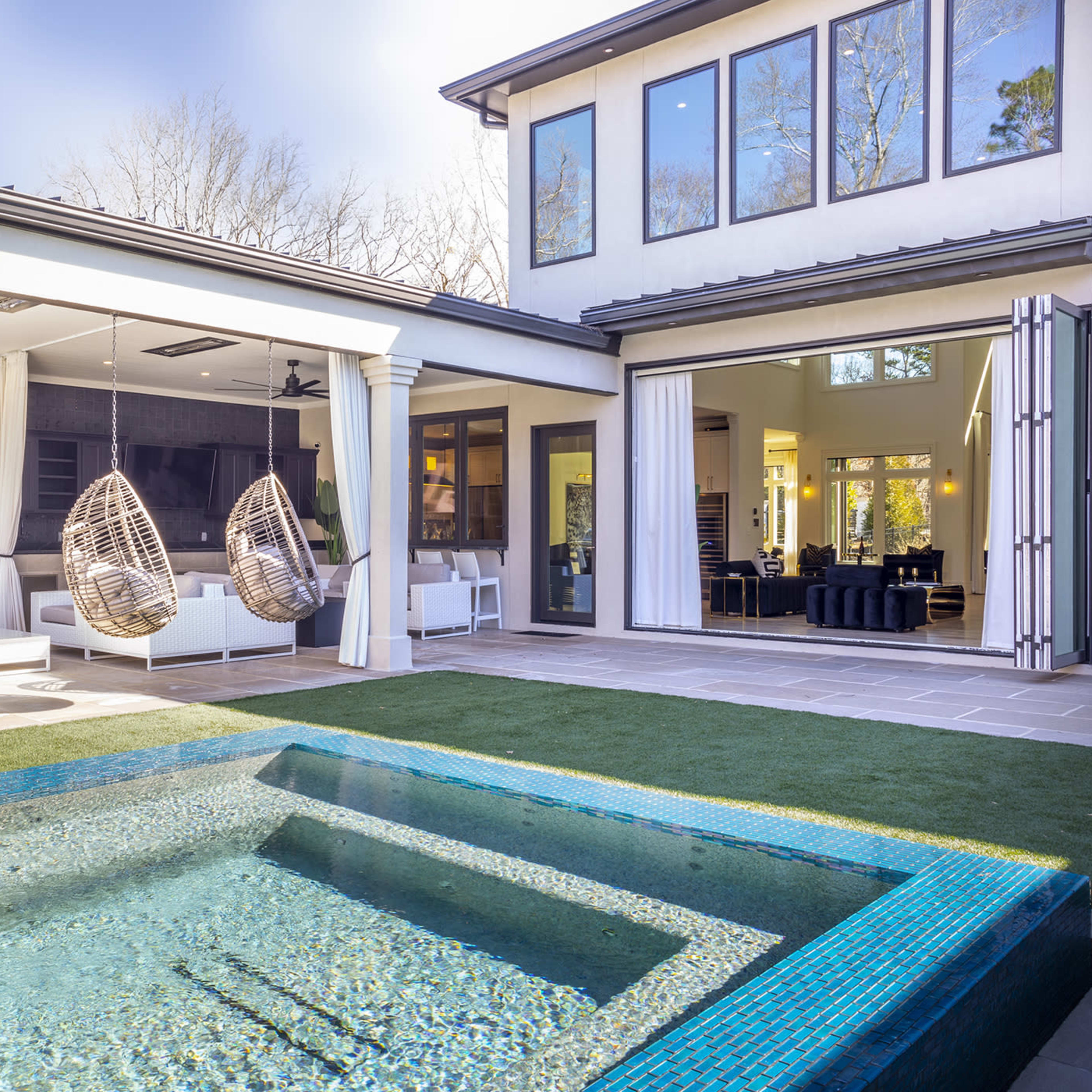 A modern backyard with a small pool, a pair of hanging chairs under a covered area, and a view of a house with large windows and an open interior space.
