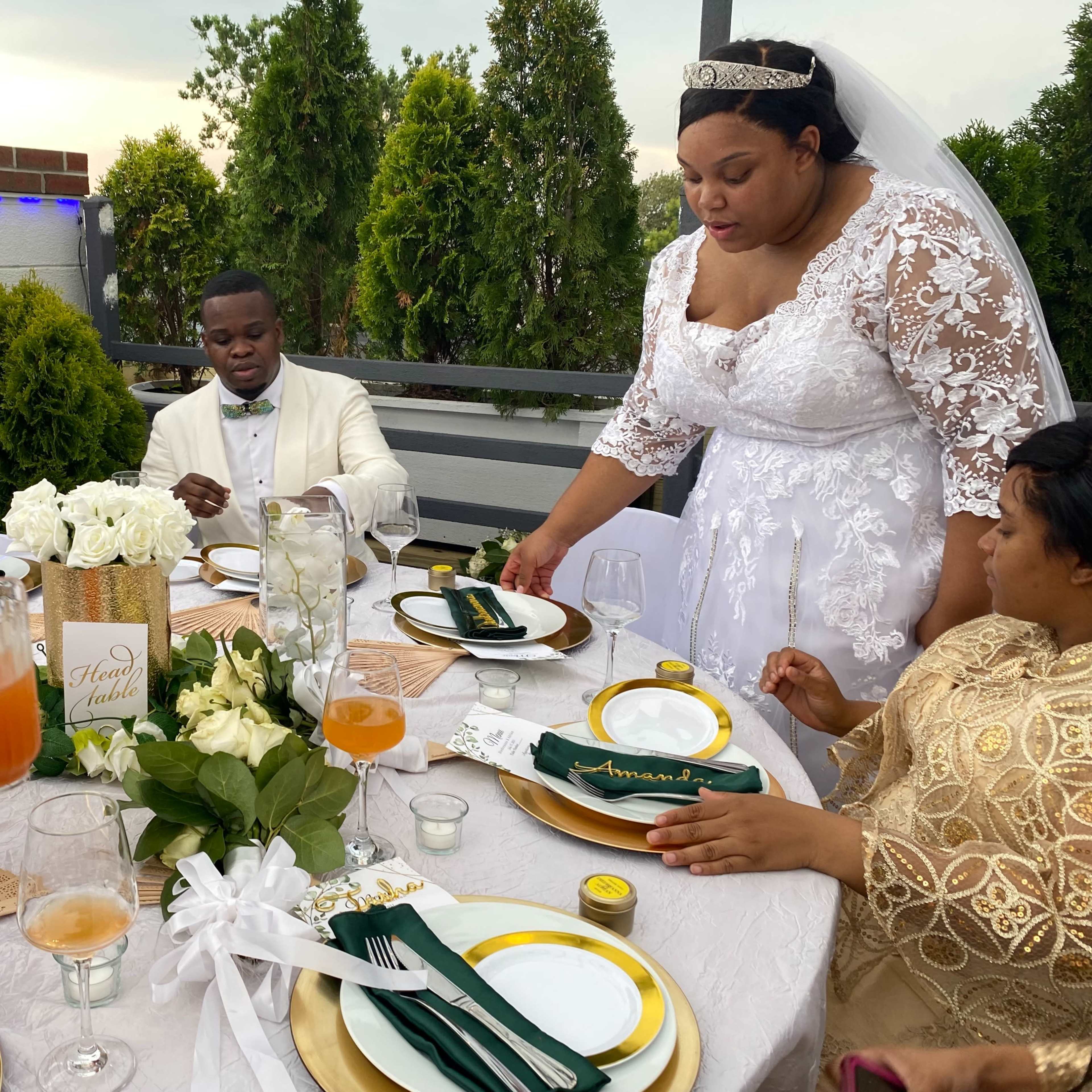 A bride in a white wedding dress serves food at a decorated outdoor reception table surrounded by guests in formal attire.