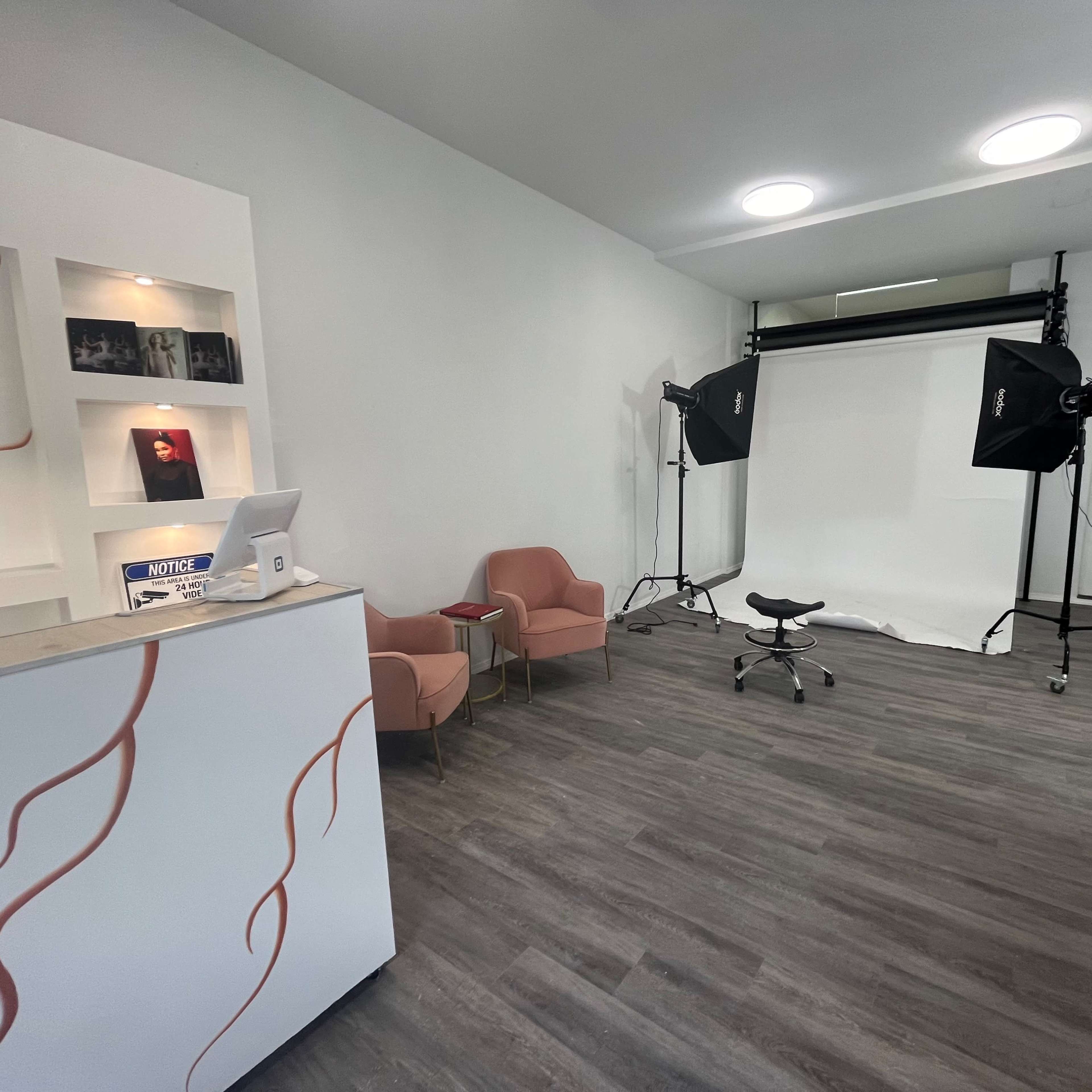A modern photo studio with a reception area featuring two pink chairs and photography equipment set up against a white backdrop.
