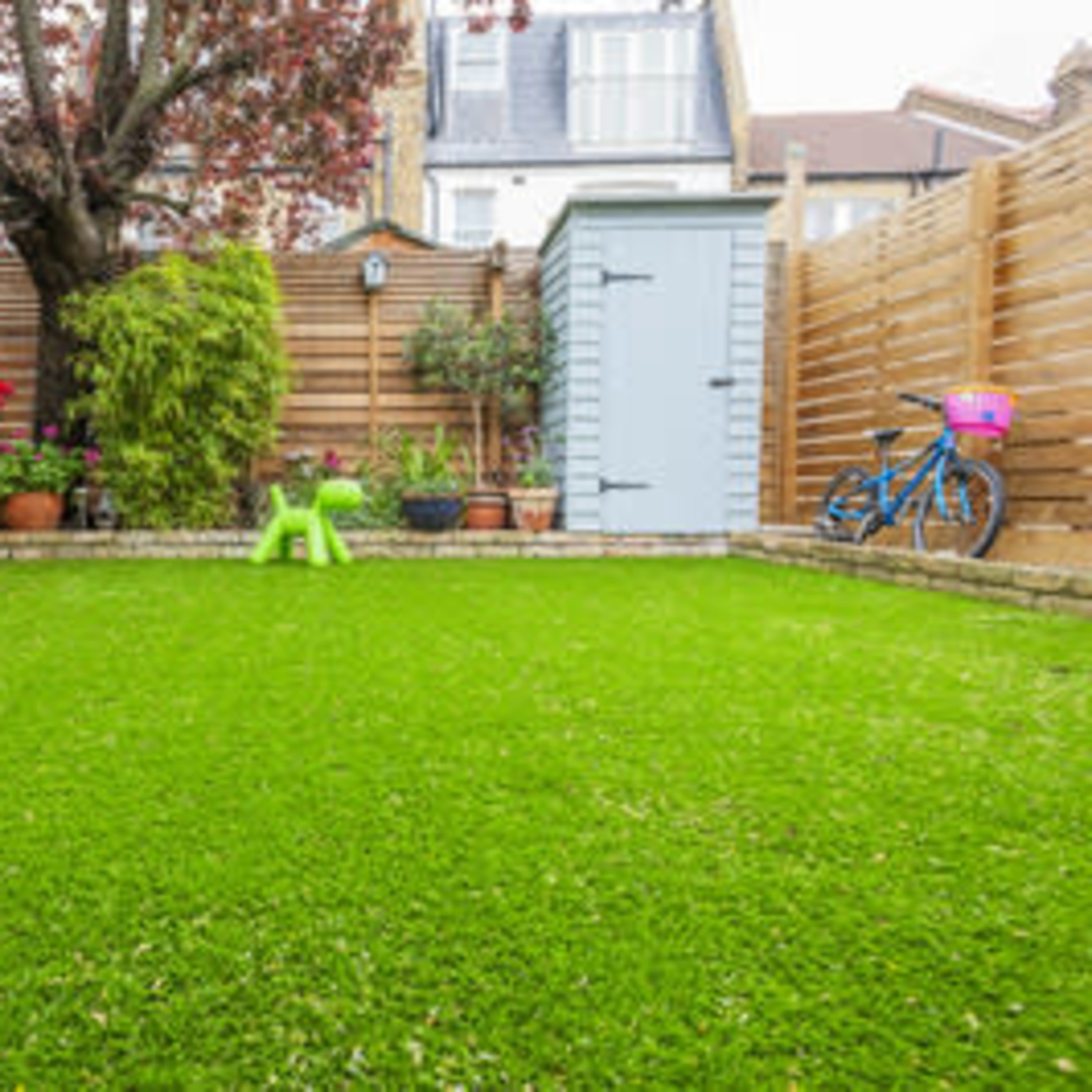 A well-maintained garden with artificial grass, bordered by wooden fencing, plants in pots, and a garden shed.