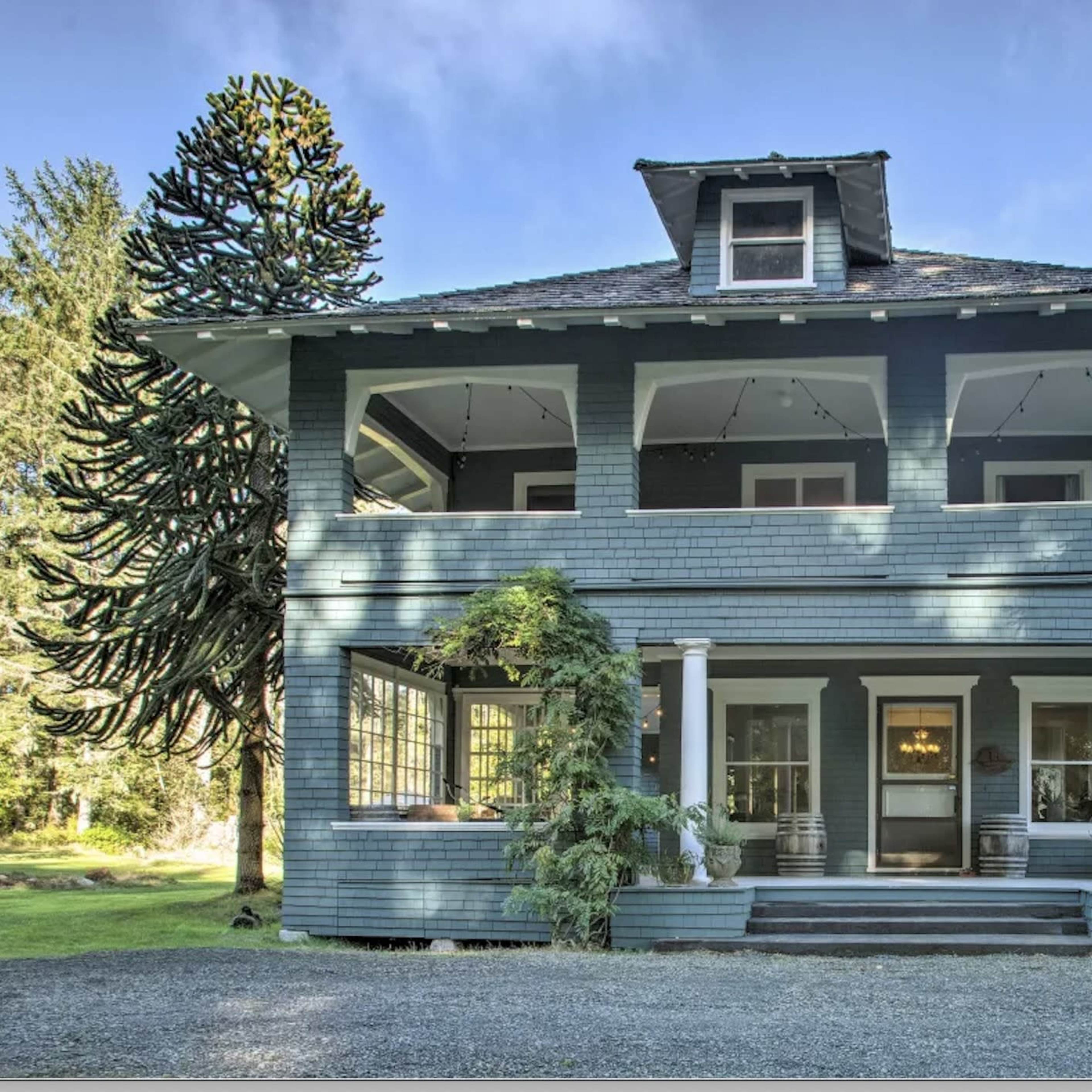 A large, two-story, blue-painted house with a covered porch, multiple windows, and an American flag hanging at the entrance.