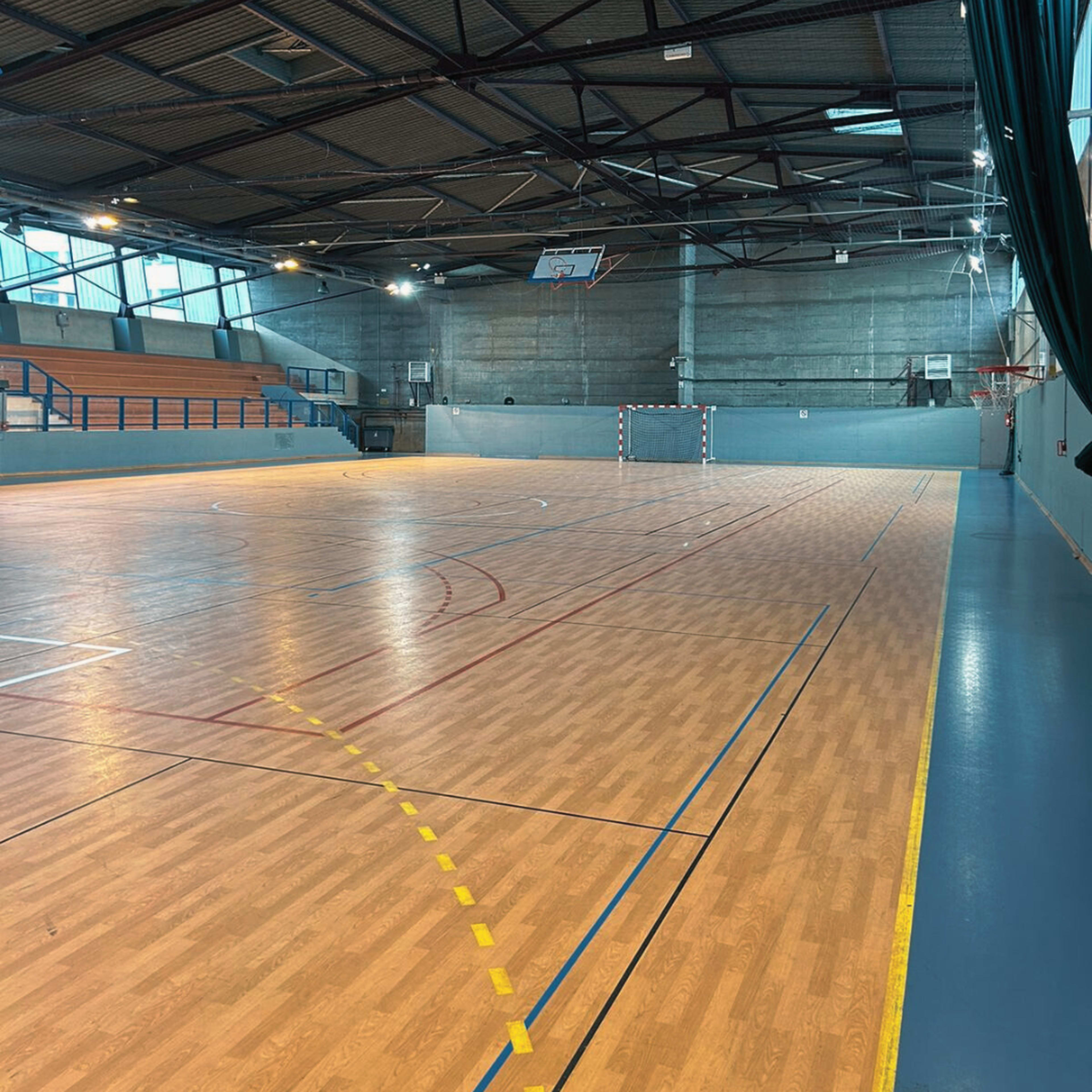 A spacious indoor sports hall with a wooden floor, basketball hoop, and goal posts.