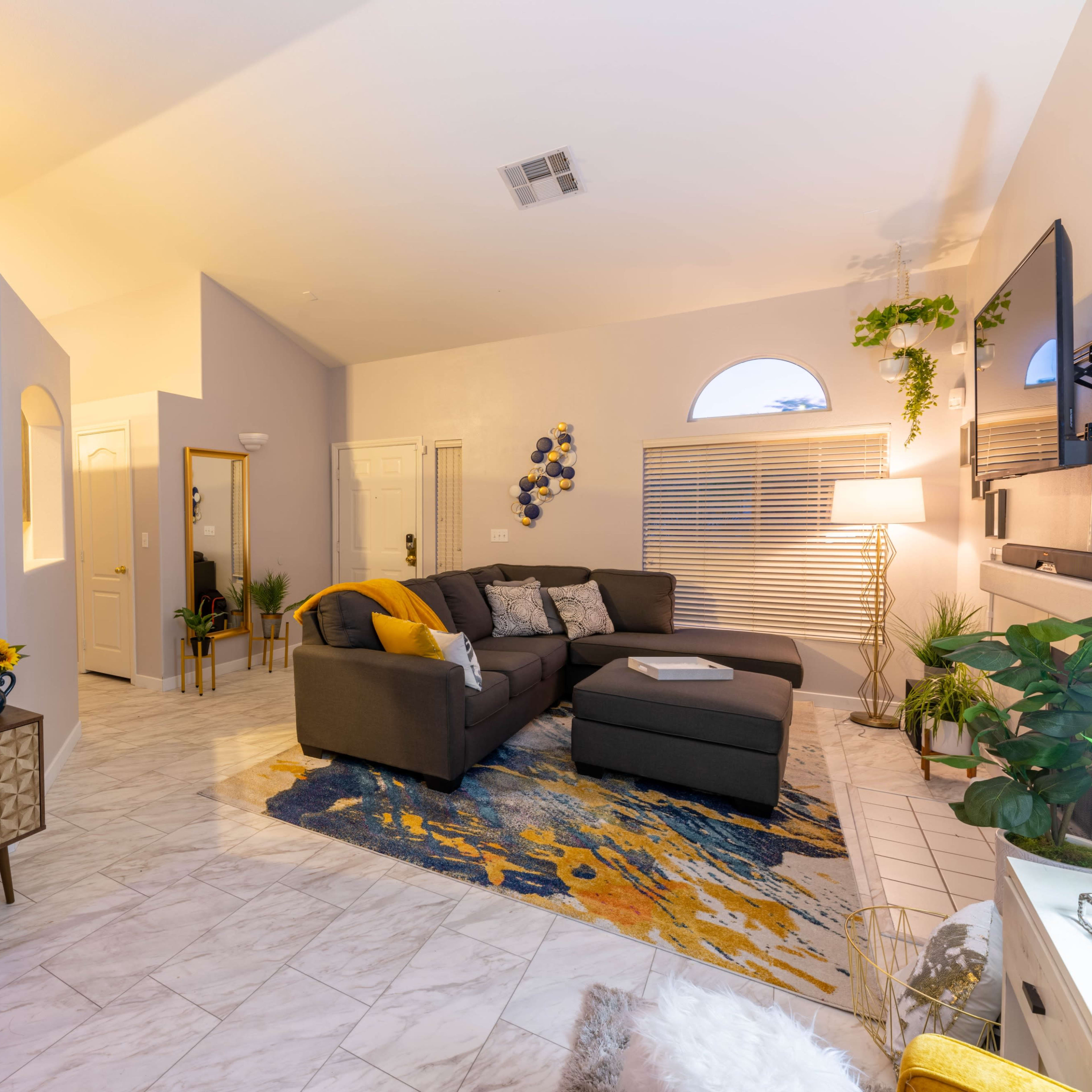 The living room features a gray sectional sofa, a colorful area rug, and a vintage-style cabinet, with a neon sign and large windows providing light.