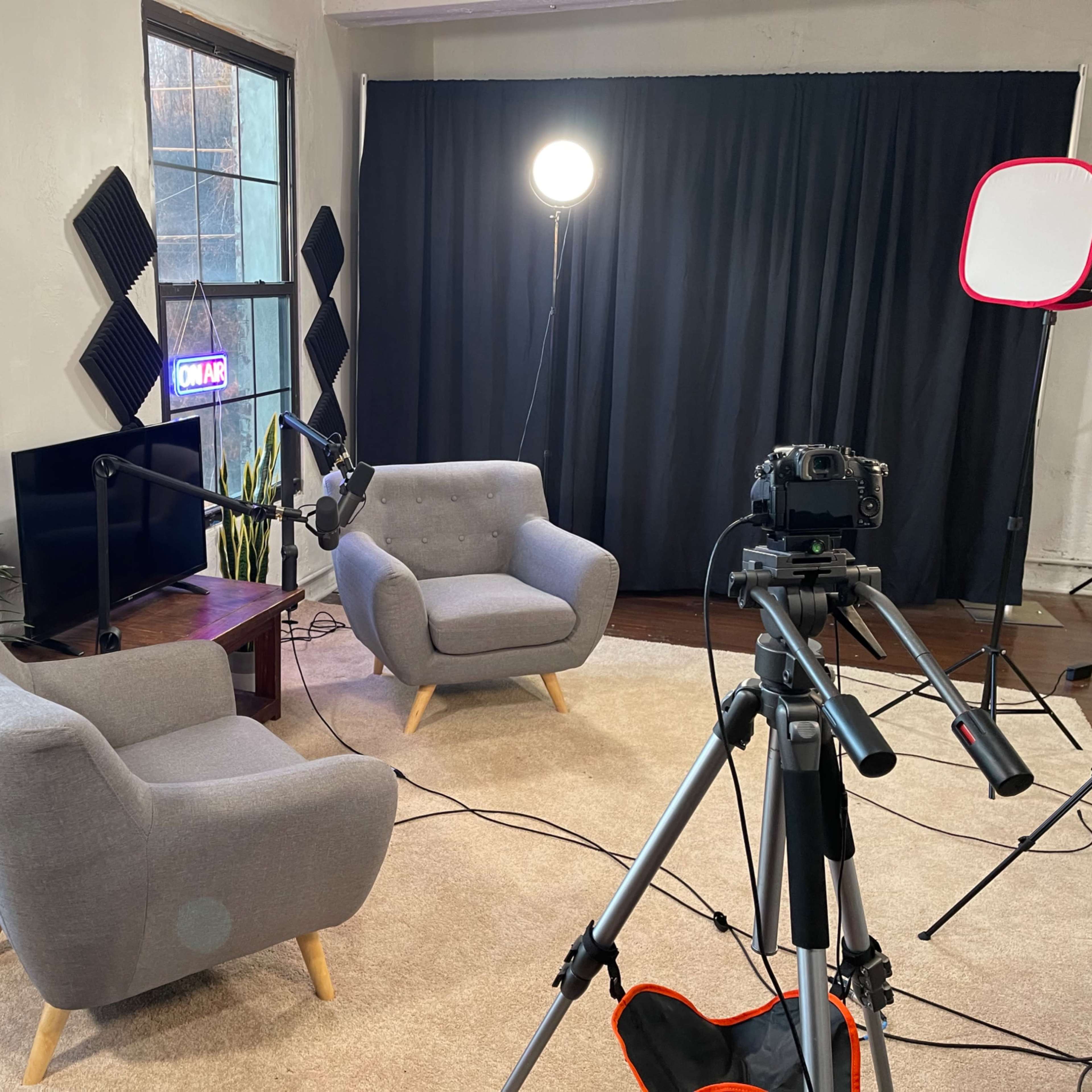 A cozy studio setup features two gray armchairs, a camera on a tripod, and a microphone, with soft lighting and black curtains in the background.