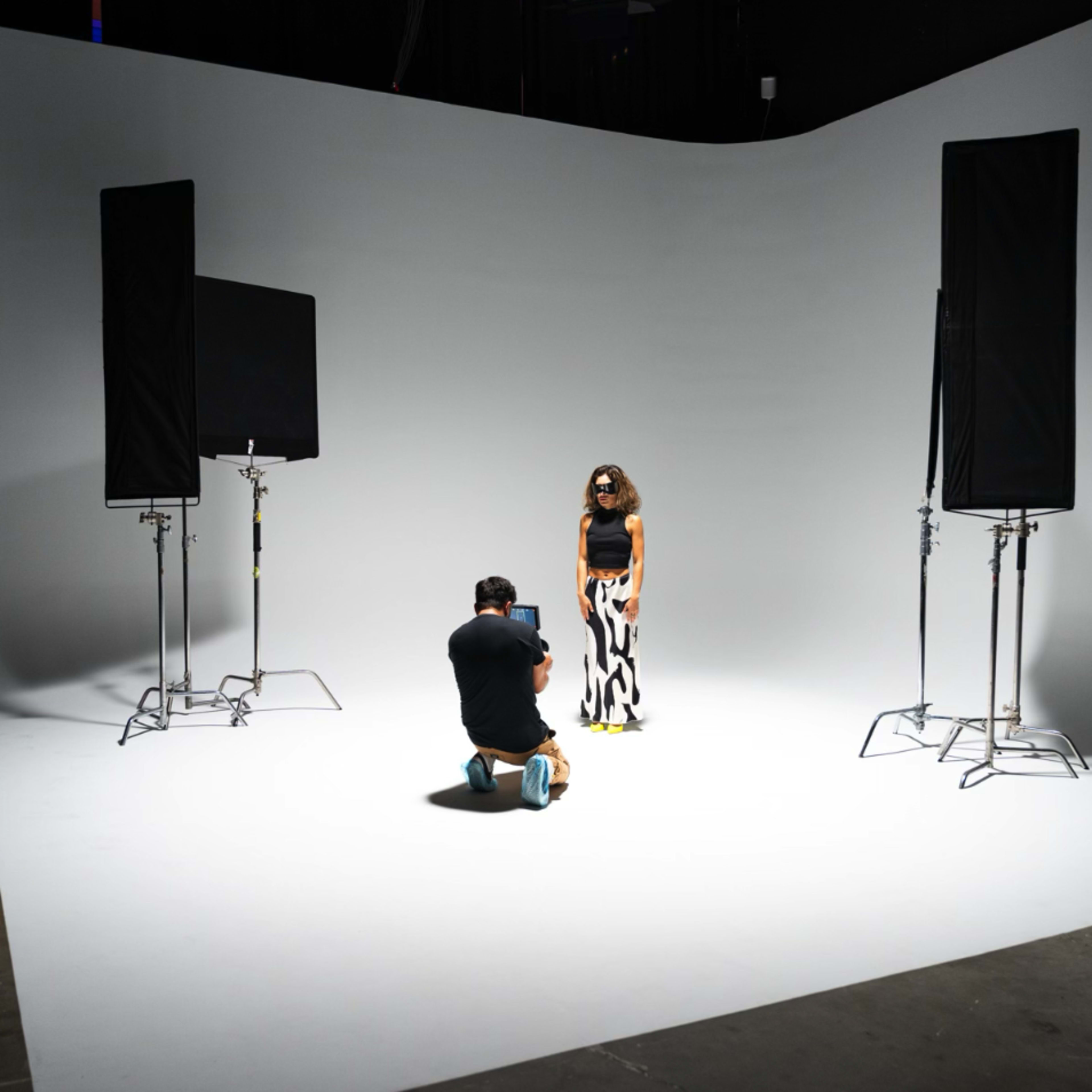 A photographer is kneeling on the floor in a studio, capturing images of a model standing on a plain white background, flanked by two lighting setups.