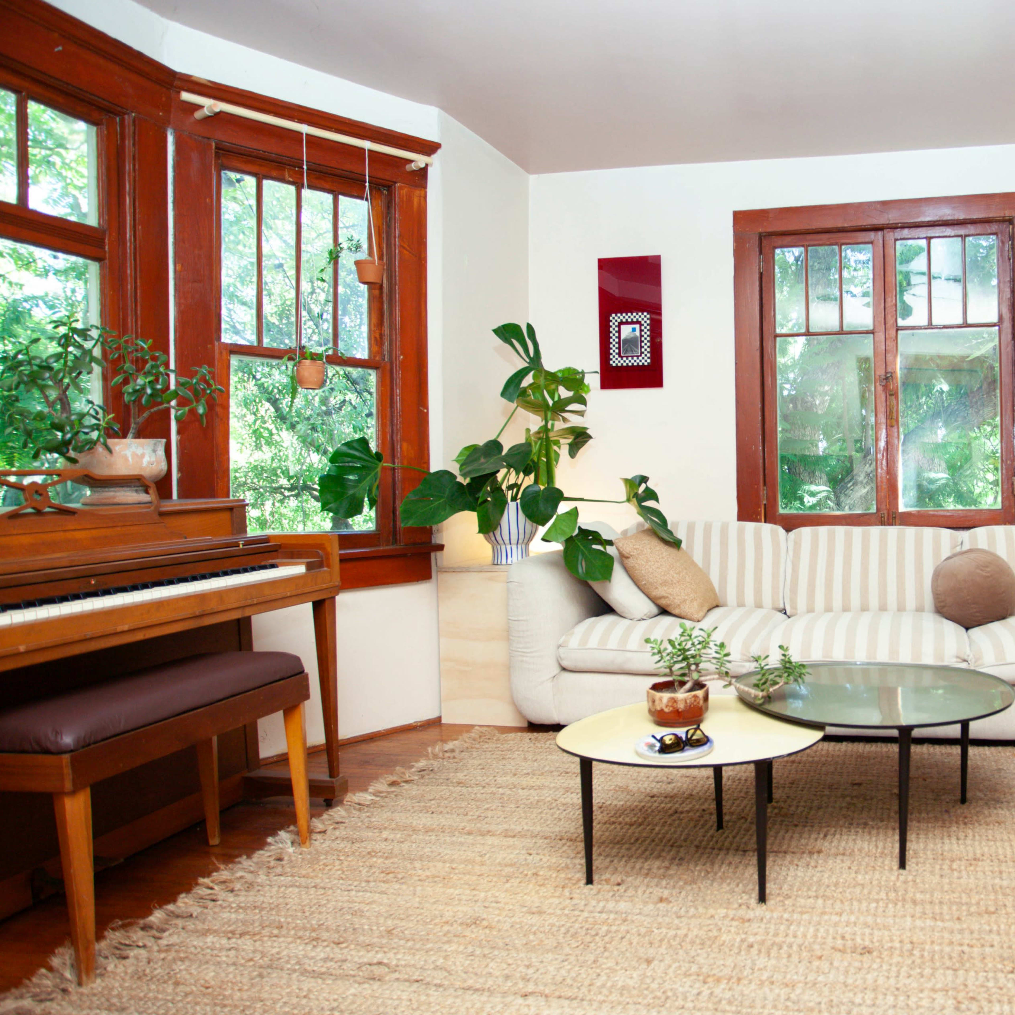 A living room featuring a striped sofa, a coffee table, a piano, and several plants, with large windows allowing natural light to enter.