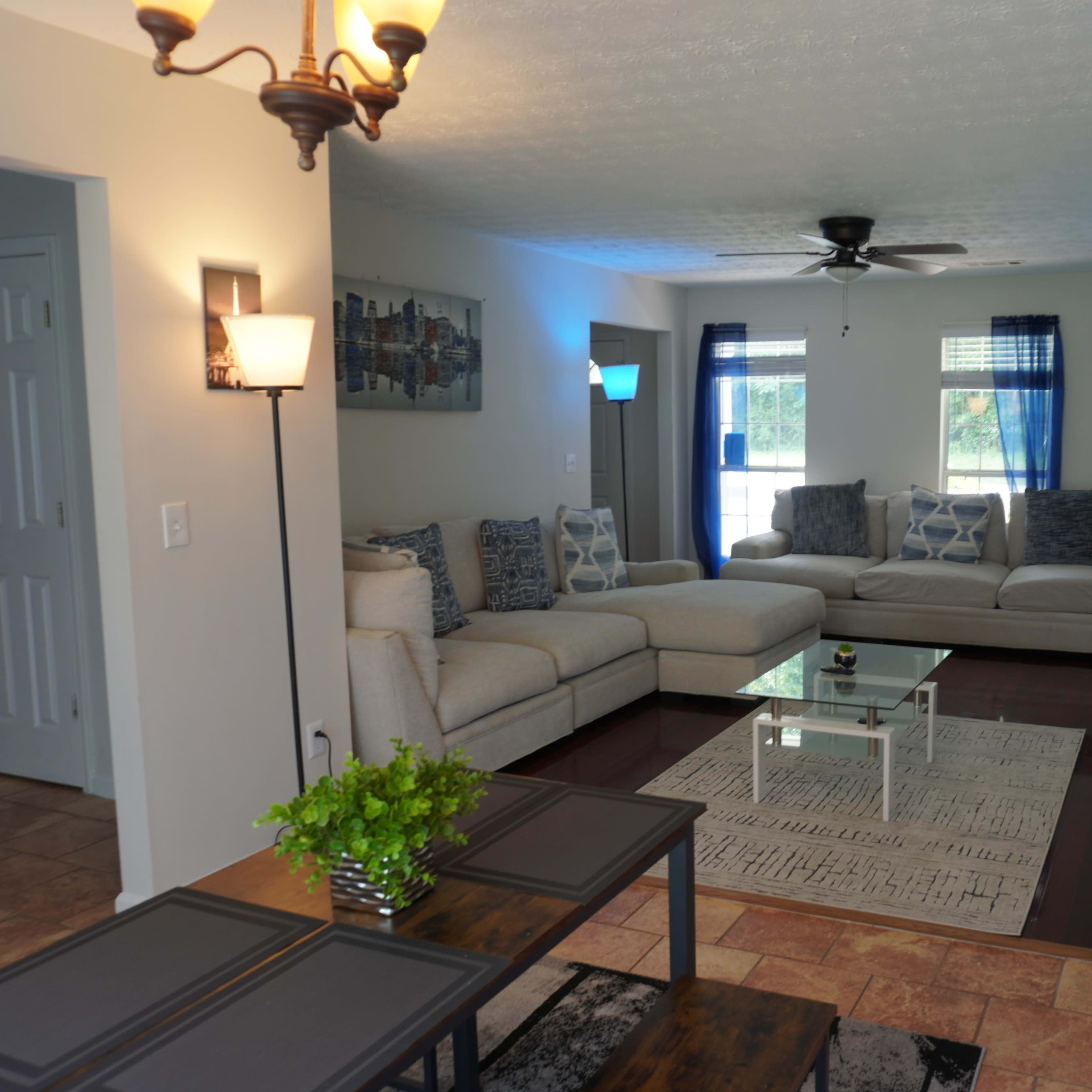 The living room features a light-colored sectional sofa, a glass coffee table, and a patterned area rug, with natural light coming through large windows.