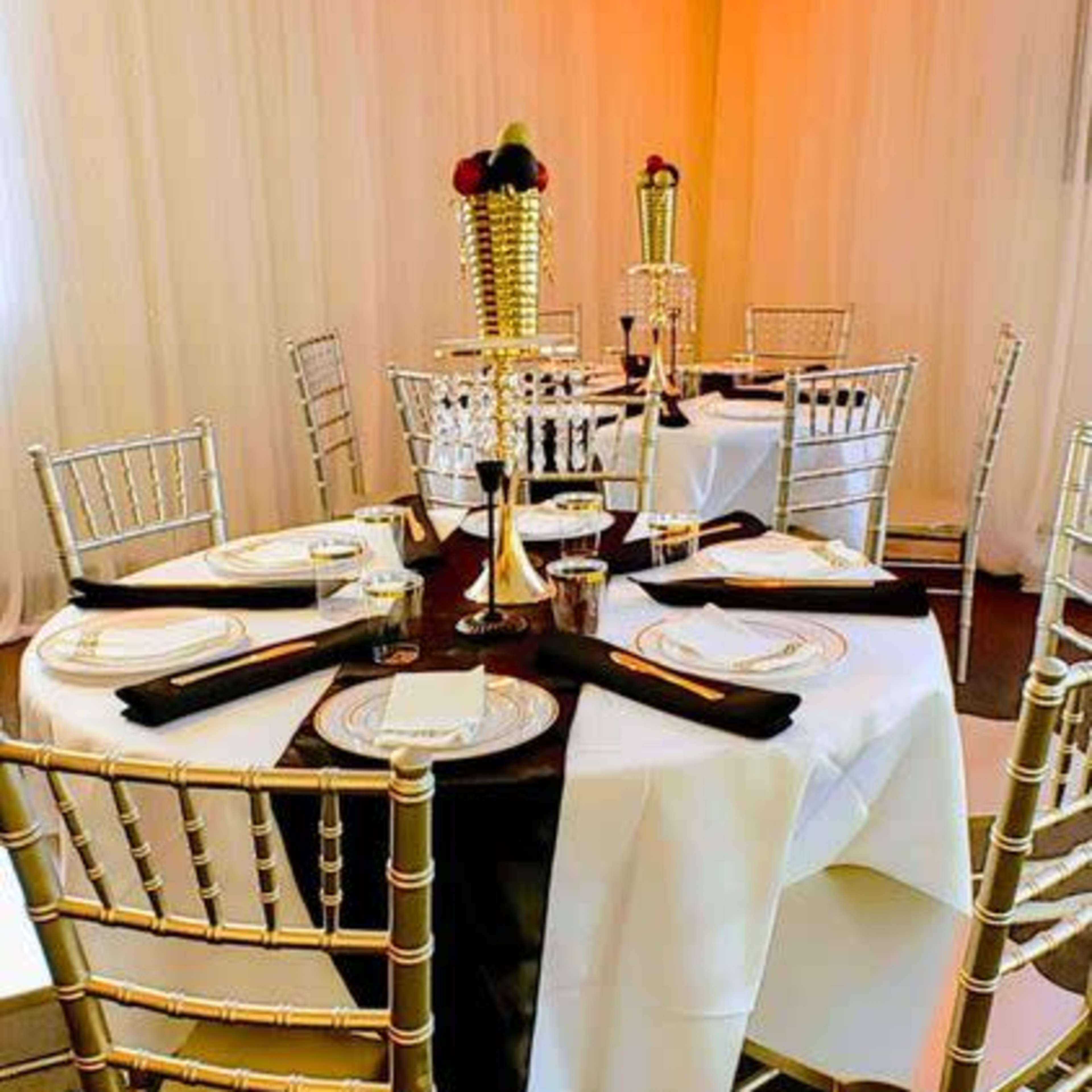 A round dining table is elegantly set with black and white table linens, surrounded by gold chiavari chairs in a softly lit event space.