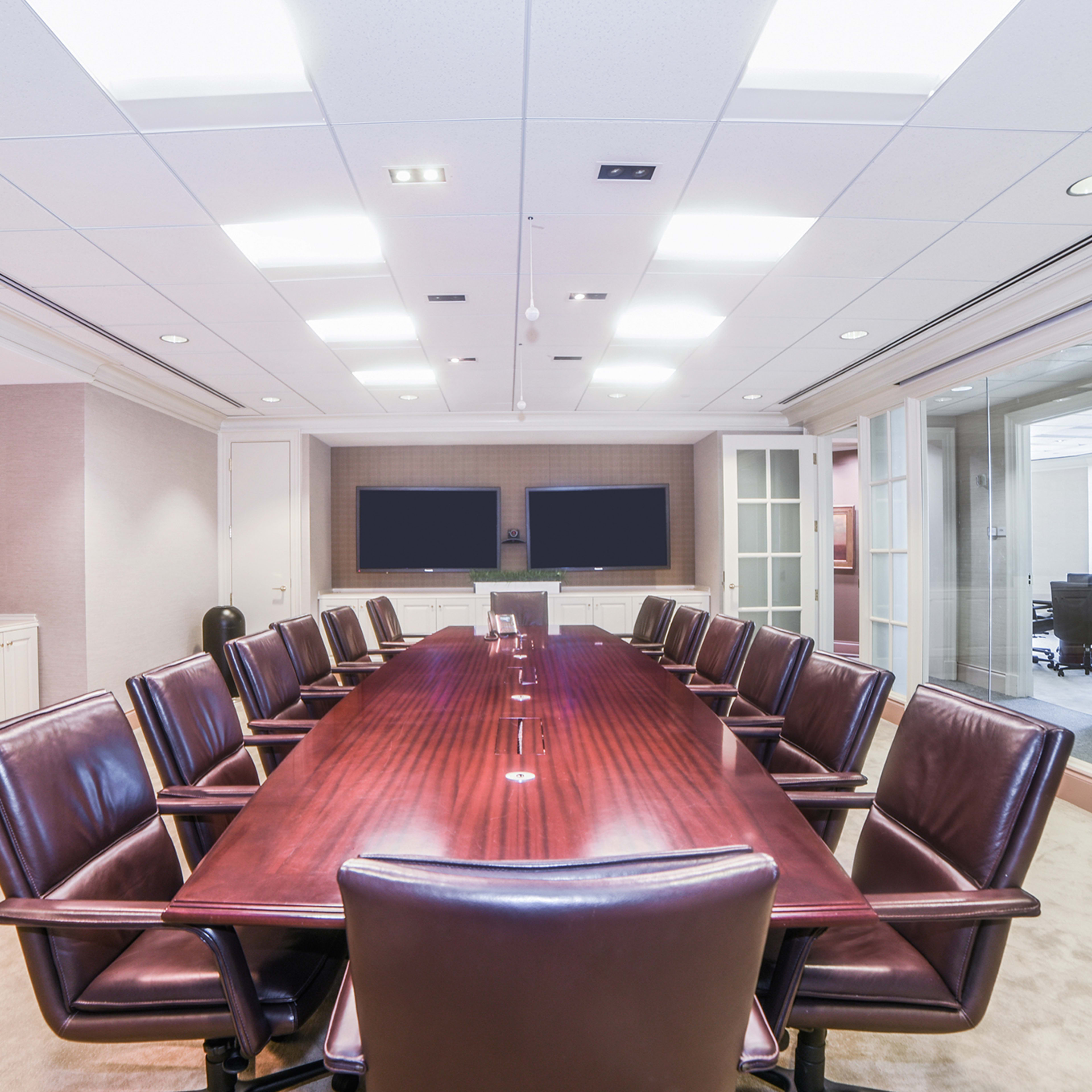 A conference room with a long wooden table surrounded by leather chairs, two large screens mounted on the wall, and glass doors leading to an adjacent office space.