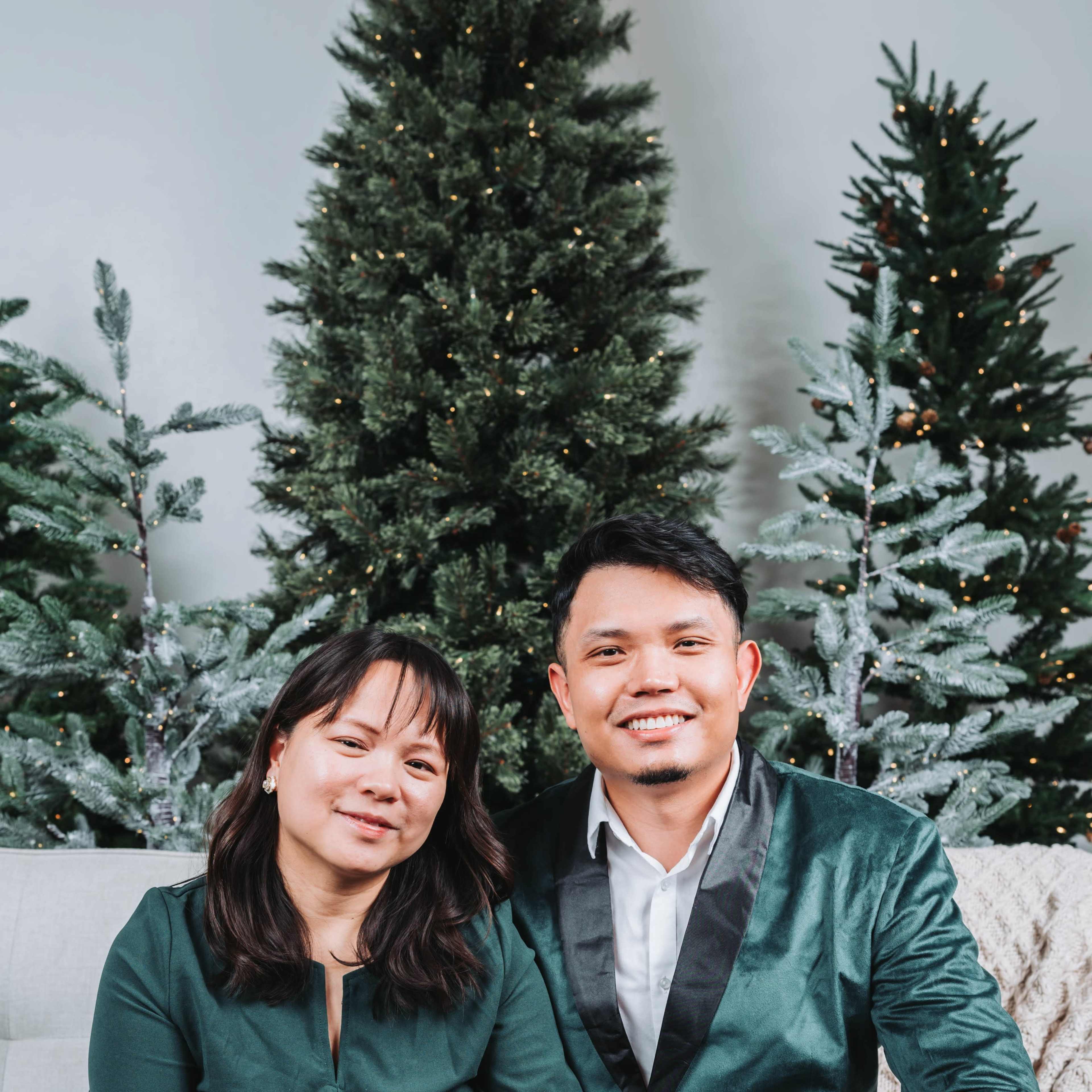 A couple sits on a sofa surrounded by festive evergreen trees decorated with lights.