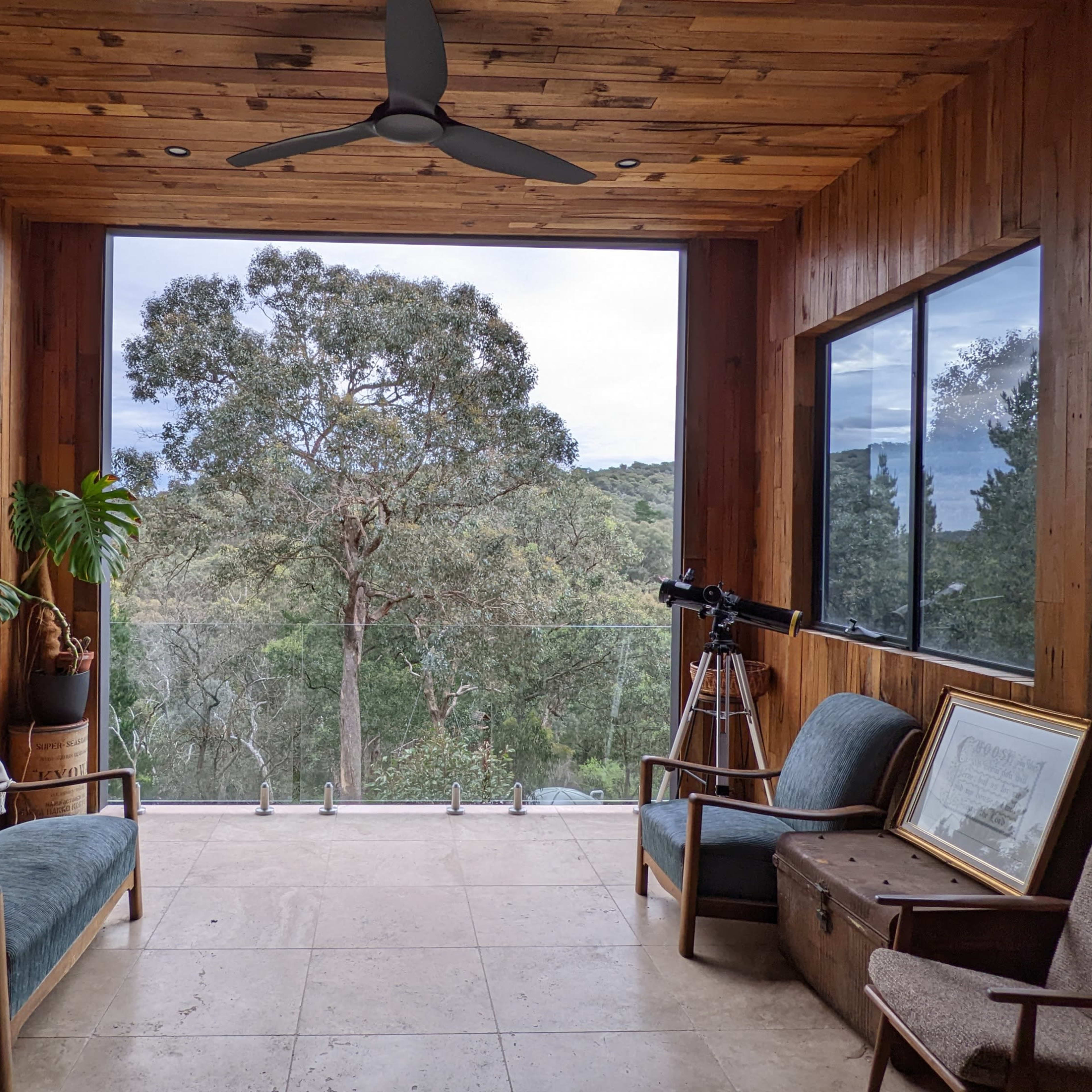 A wooden-paneled room with large windows overlooking a natural landscape, featuring two couches, a telescope, and a few pieces of furniture.