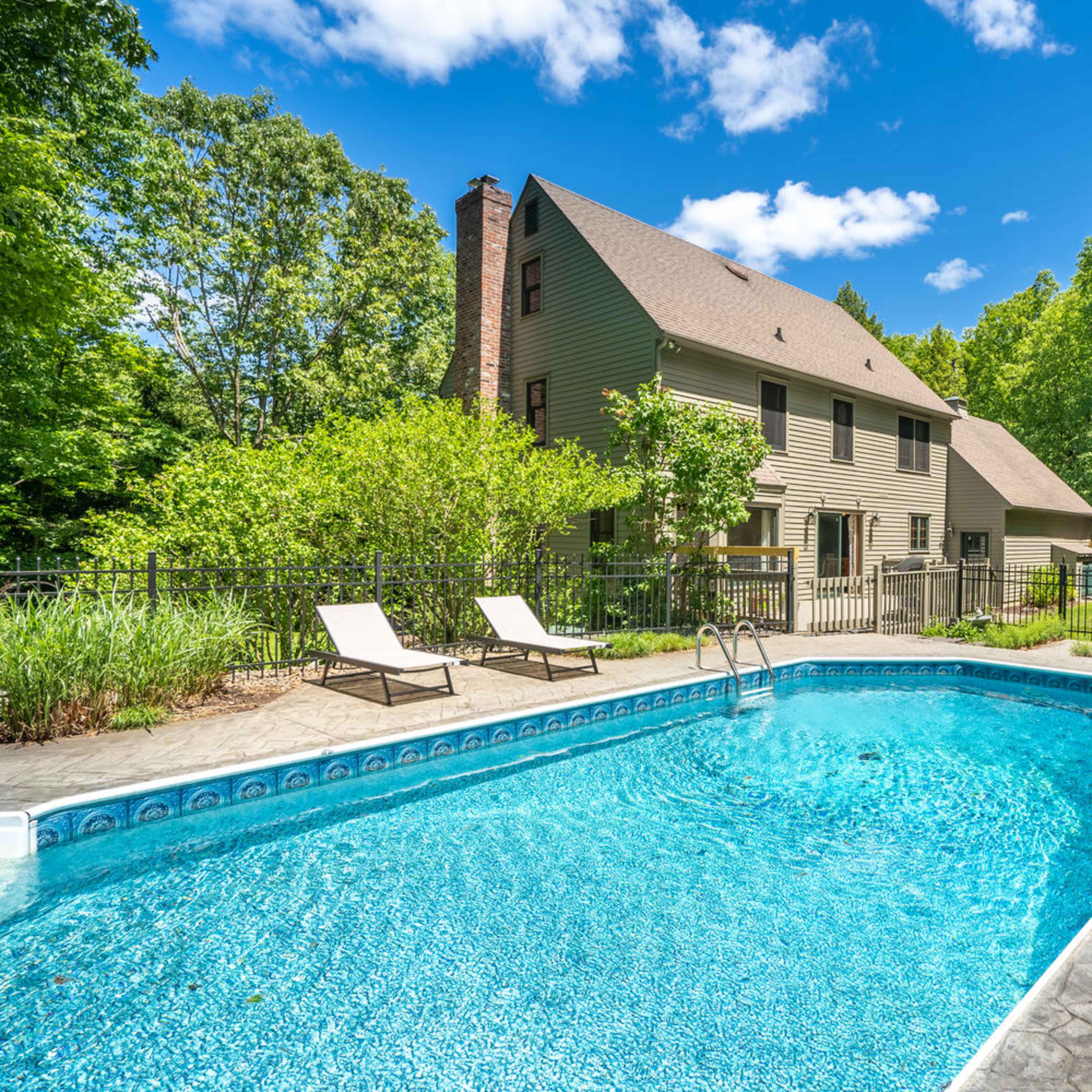 A rectangular swimming pool surrounded by a stone patio and two lounge chairs, adjacent to a two-story house, all set against a backdrop of lush green trees.