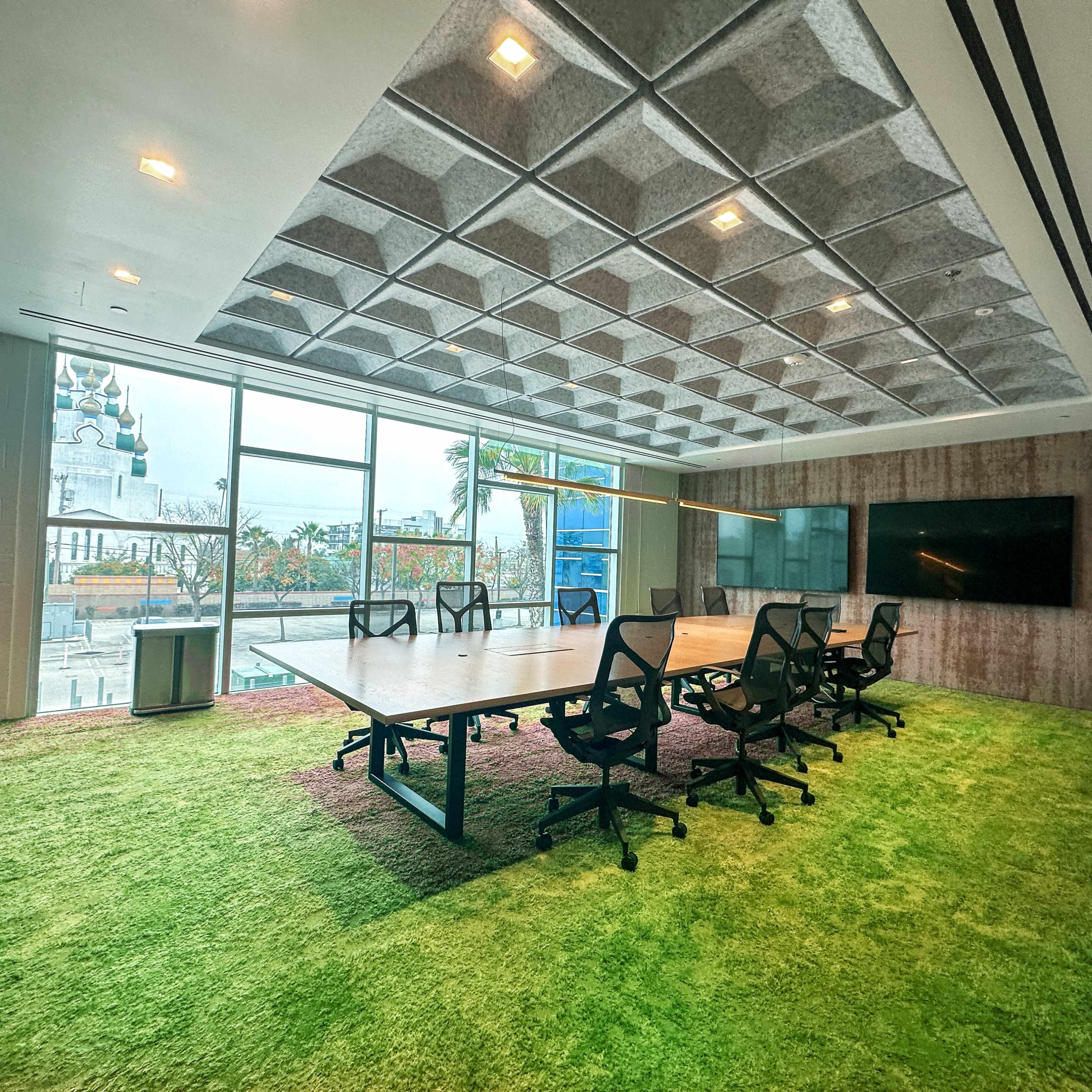 A modern conference room features a large table surrounded by ergonomic chairs, with green carpet and wide windows that overlook an outdoor area.
