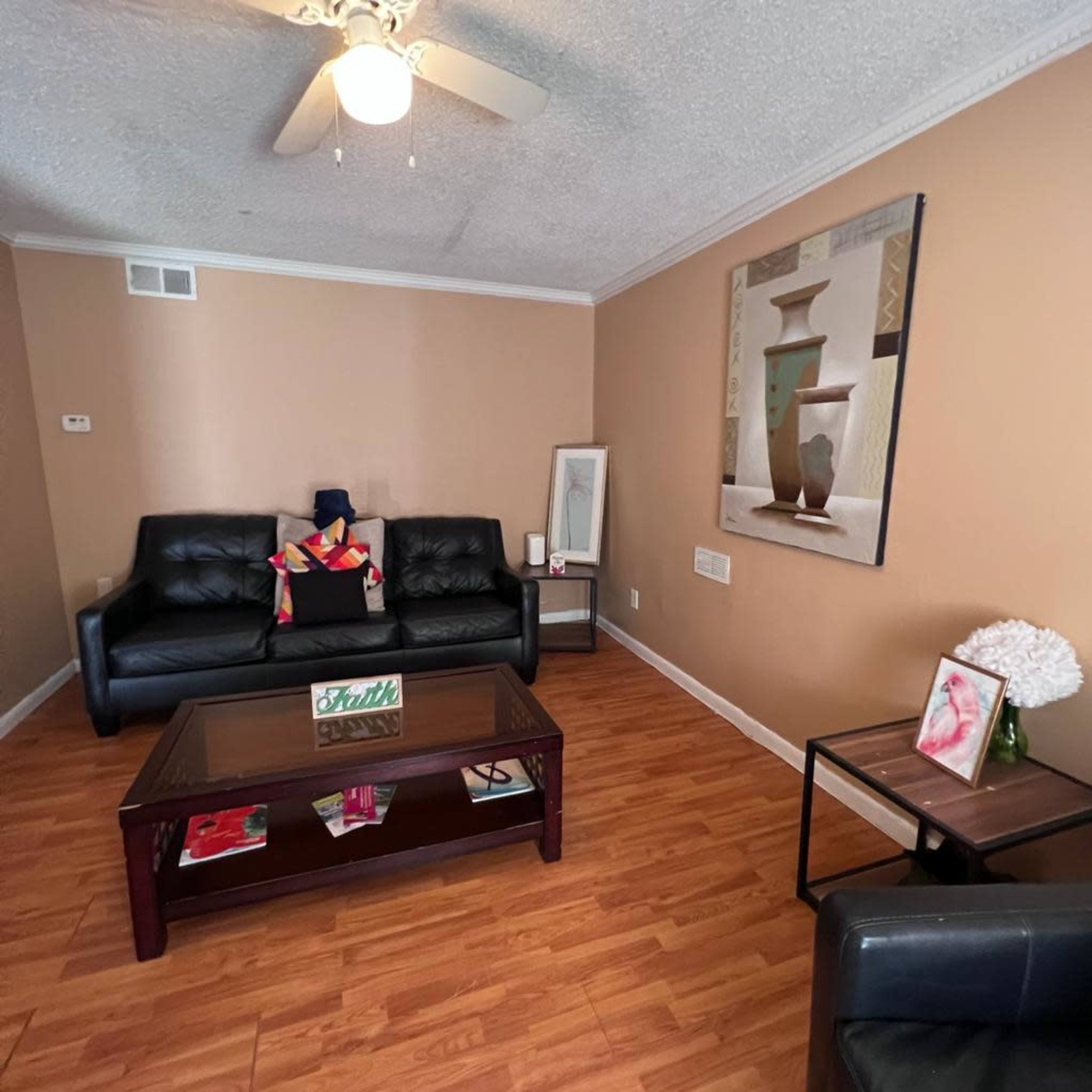 The room features a brown-painted wall with a black leather sofa and a glass coffee table, complemented by a ceiling fan and decorative art on the wall.