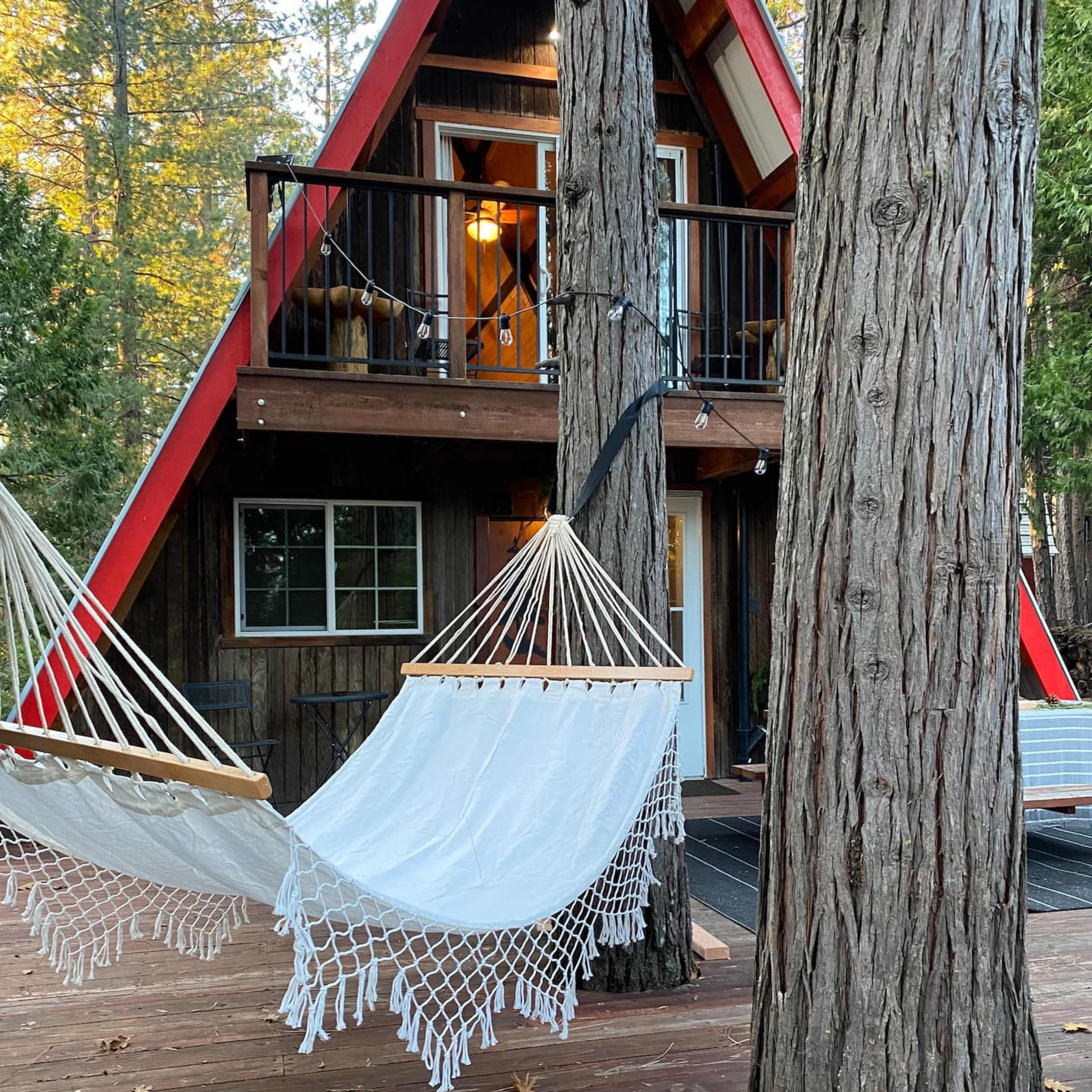 A quaint A-frame cabin with red accents is situated among tall trees, featuring two hanging hammocks on a wooden deck.