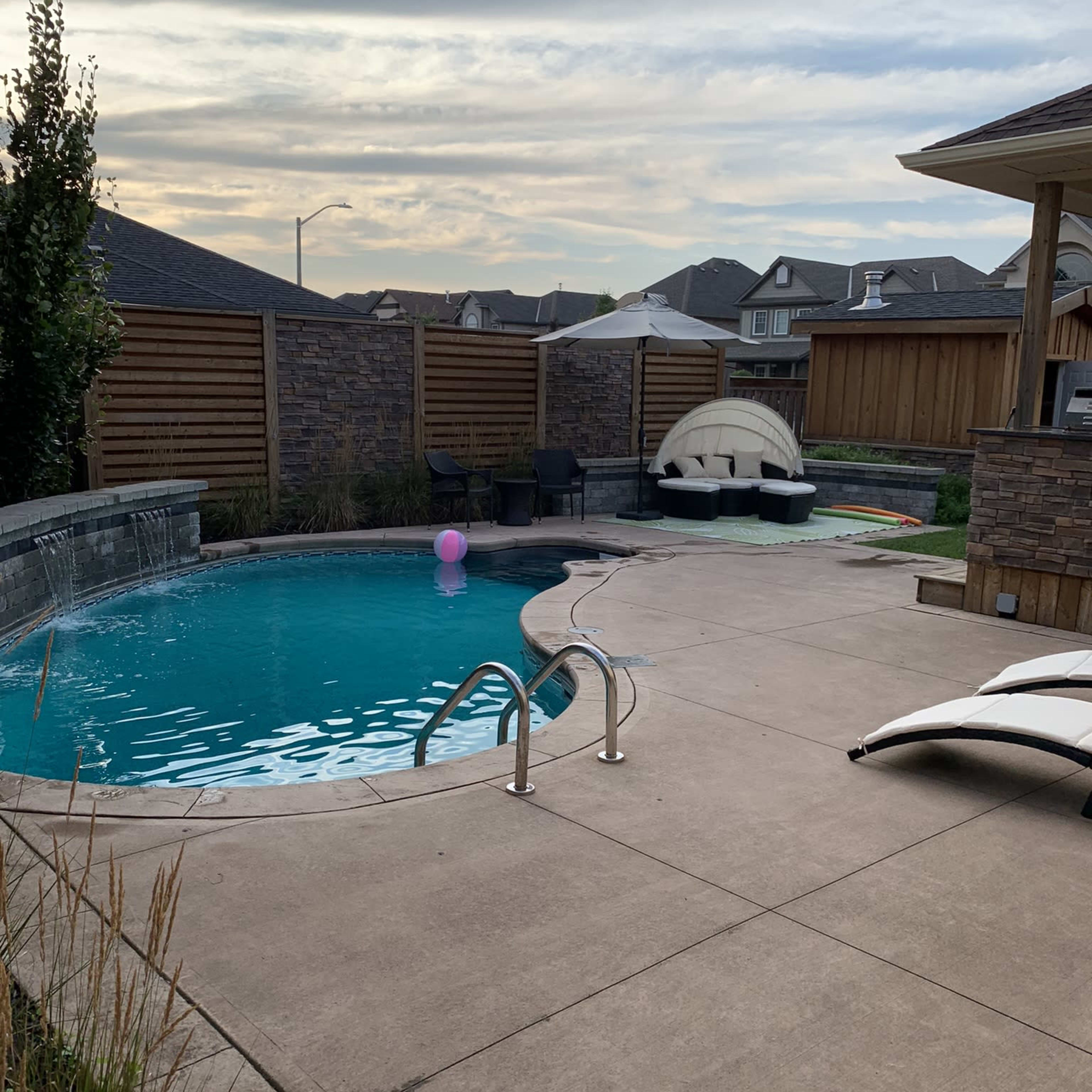 A pool area with a blue swimming pool, a slide, and lounge chairs on a paved patio surrounded by wooden fencing and landscaping.