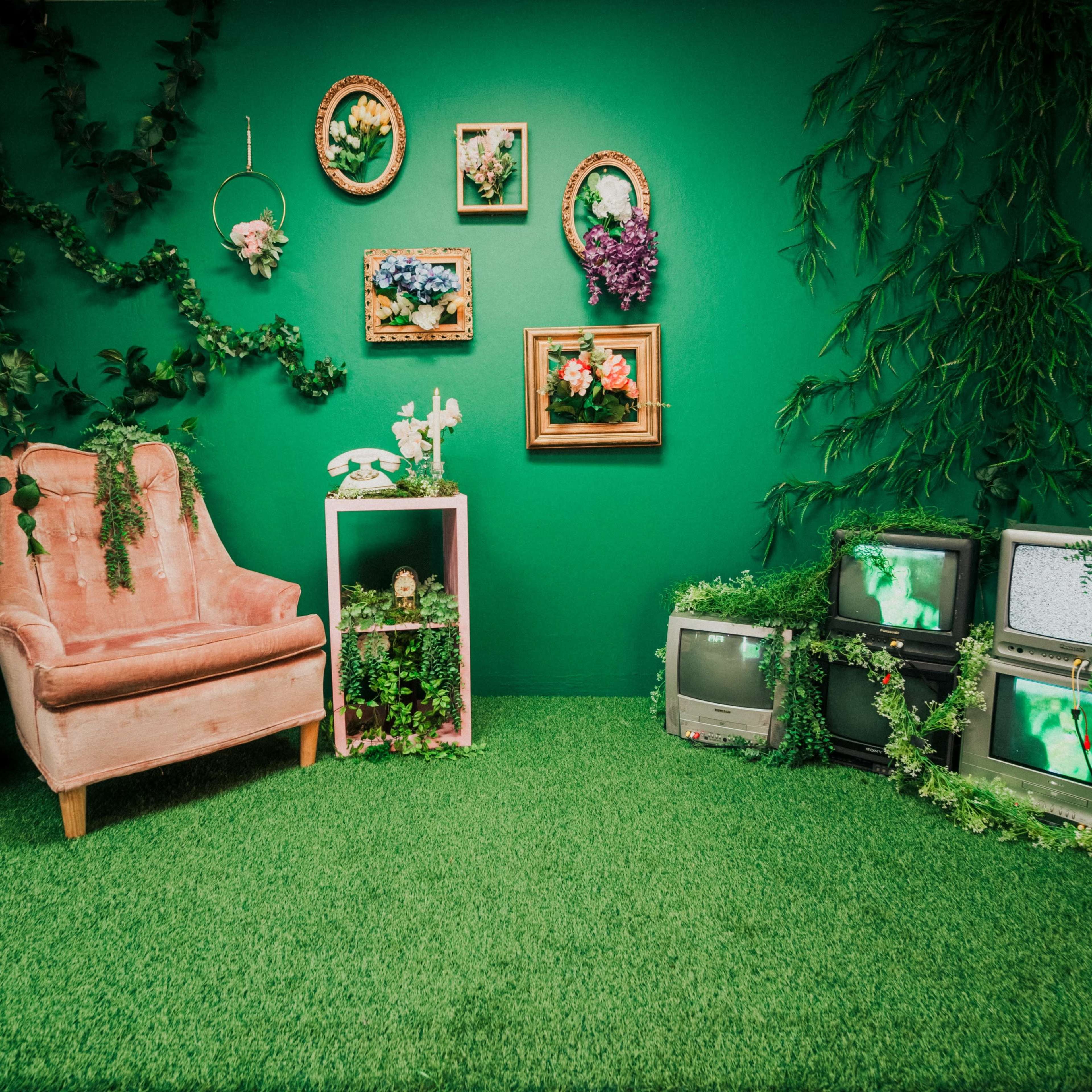 A pink upholstered chair sits against a vibrant green wall adorned with framed artwork and vintage televisions surrounded by decorative plants.