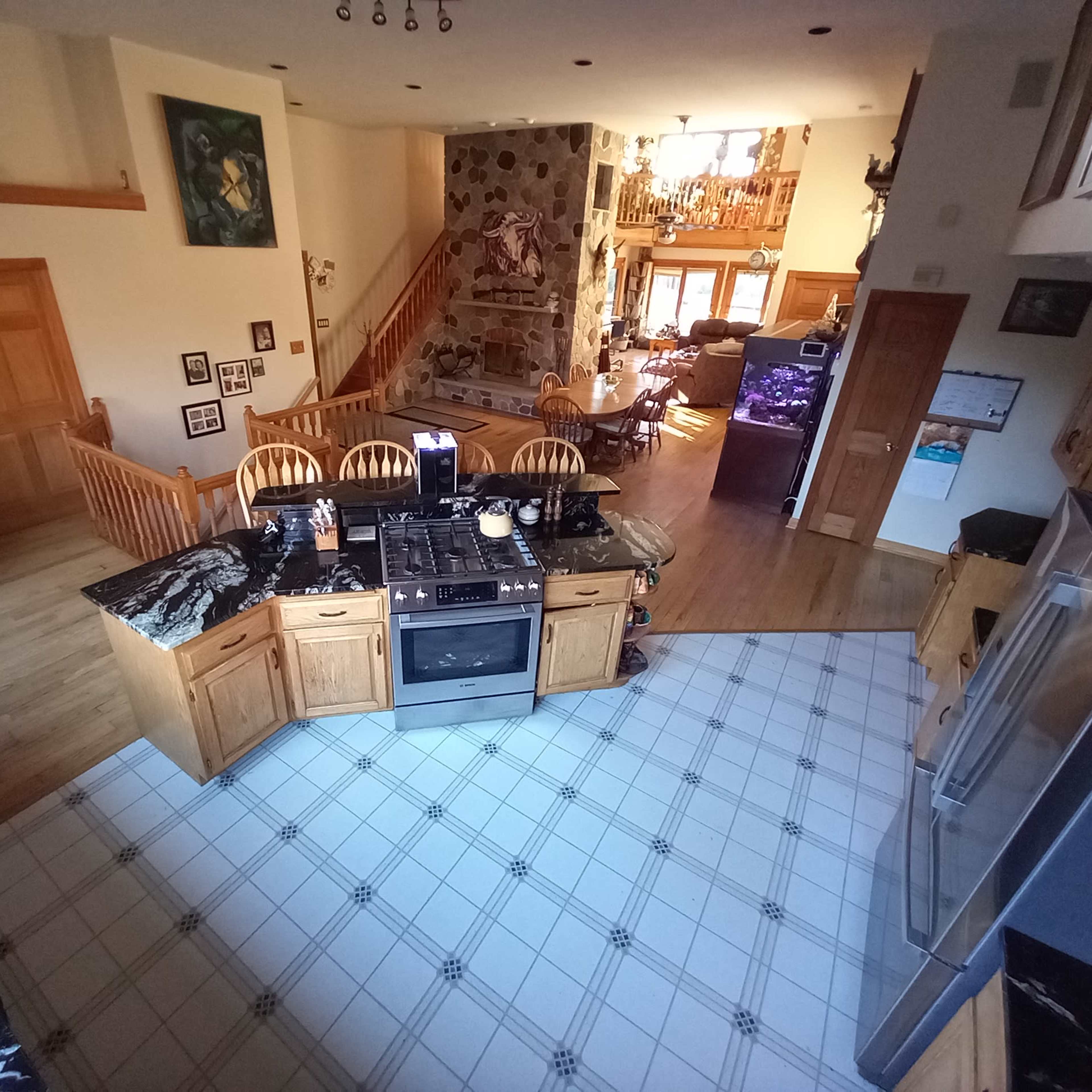 A spacious kitchen with a black and white tiled floor, wooden cabinets, and a central island, overlooking a bright living area with a staircase and a stone wall.