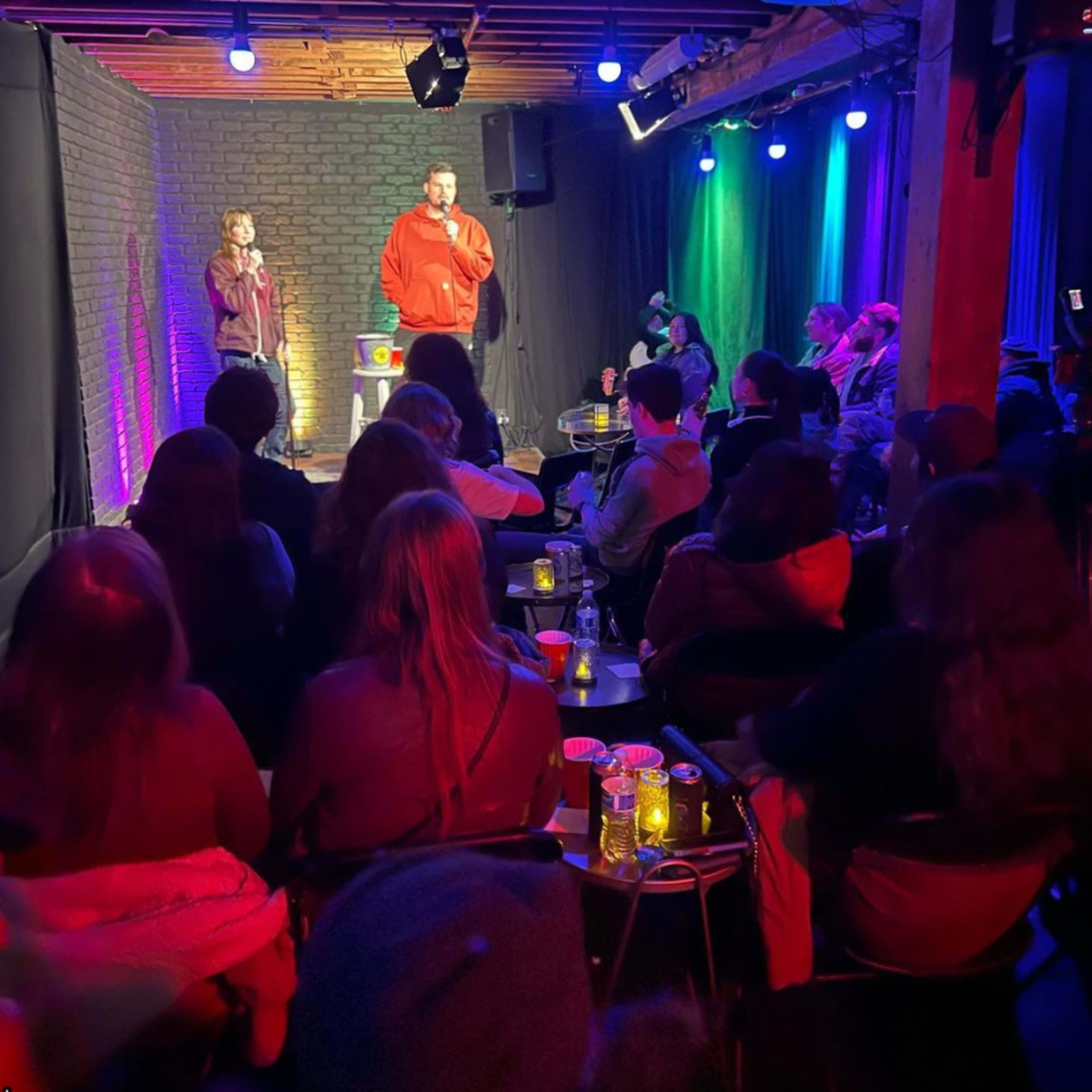 A comedian performs on stage in a cozy venue while an audience watches attentively from their seats.