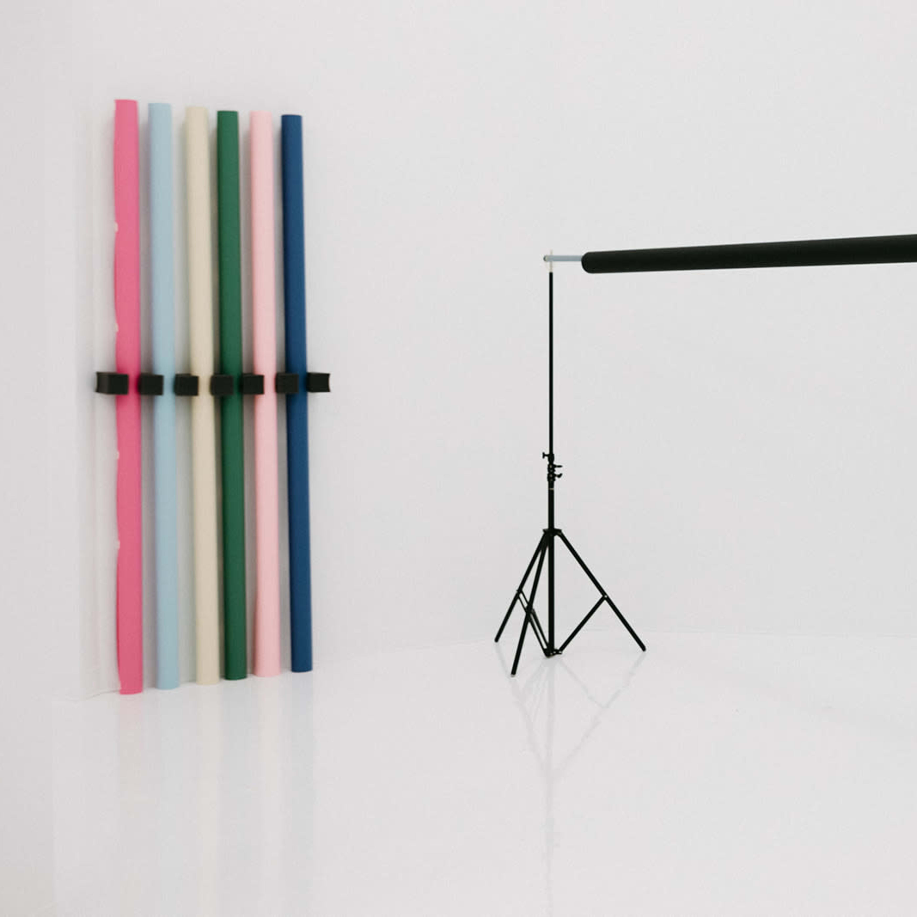 A photography studio with two black backdrop stands and a roll of black paper suspended between them, alongside a vertical arrangement of colorful paper rolls on the wall.