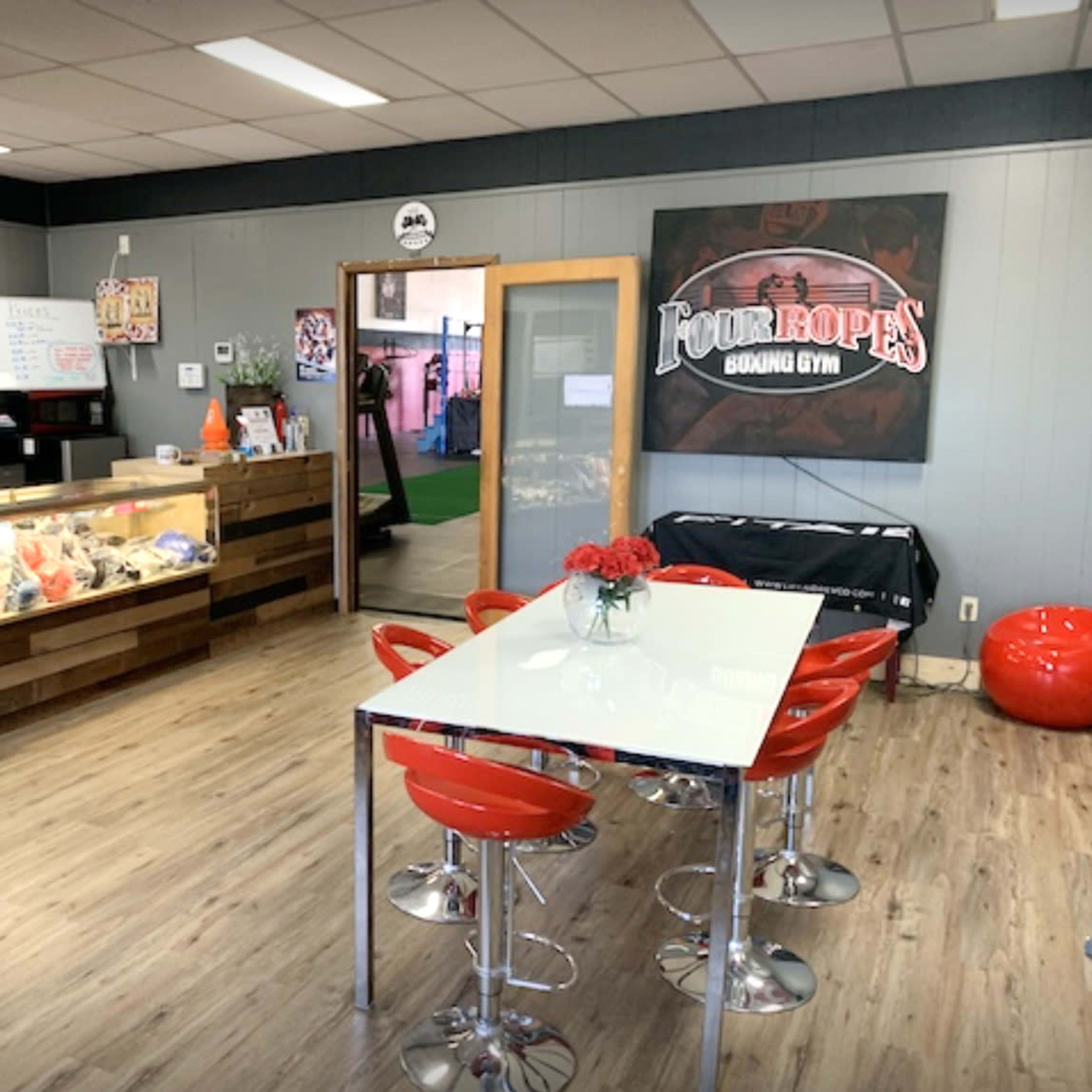 The interior of a boxing gym's reception area, featuring a front desk, a display case, a dining table with red chairs, and fitness-related decor on the walls.