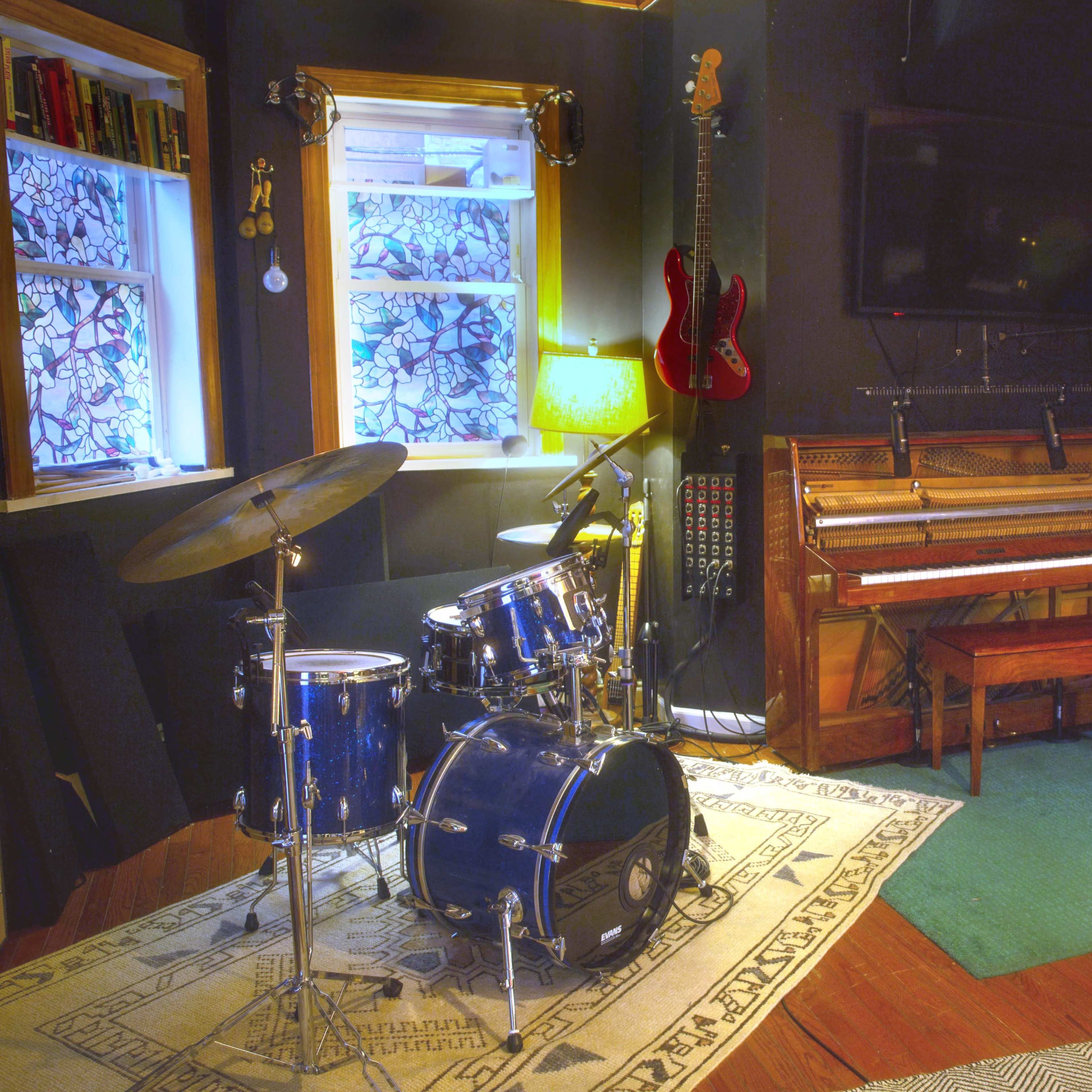 A music studio featuring a drum set, an acoustic piano, and electronic keyboards arranged against a dark wall, with windows providing natural light.