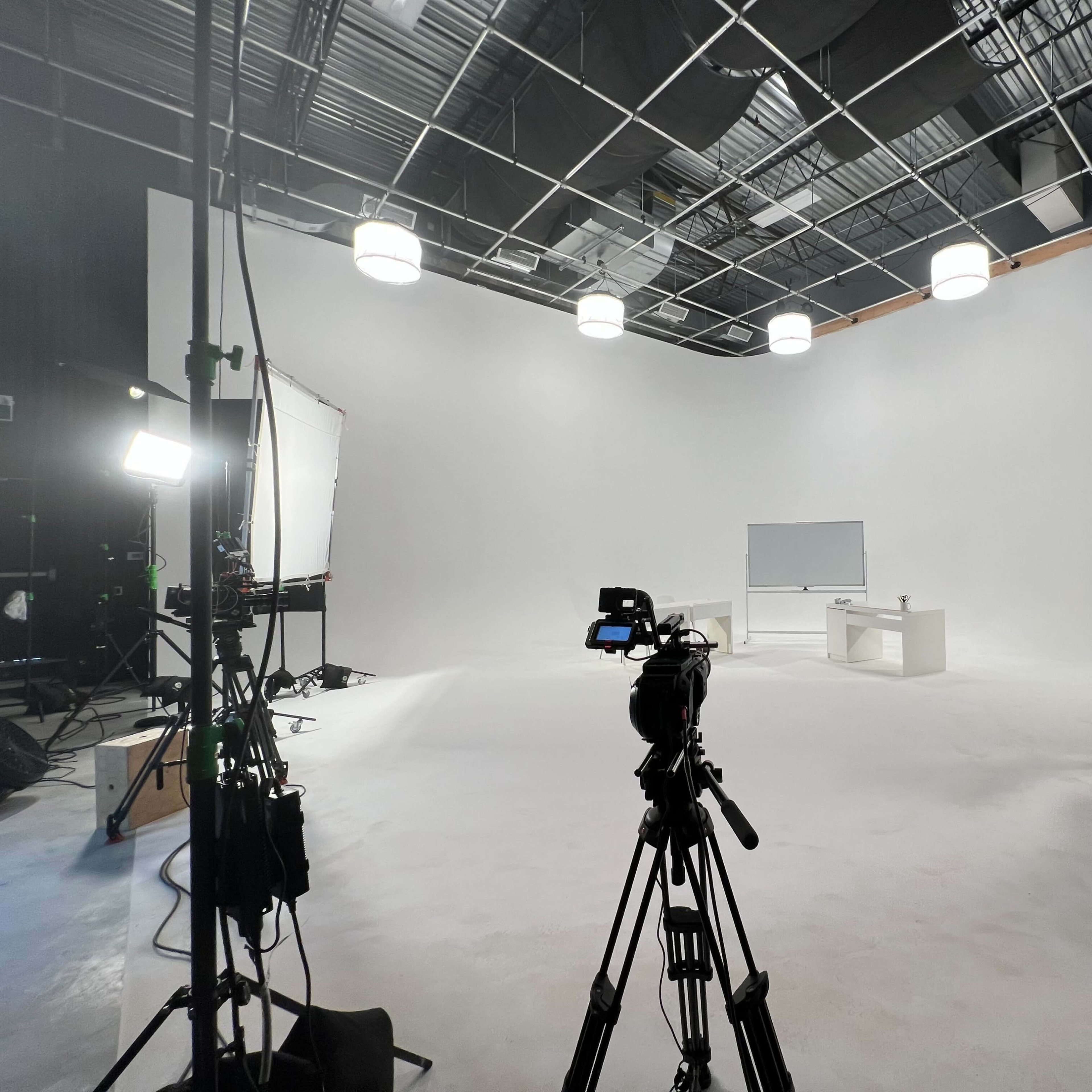 A film studio setup features a white backdrop, two cameras on tripods, a whiteboard, and several lighting fixtures.