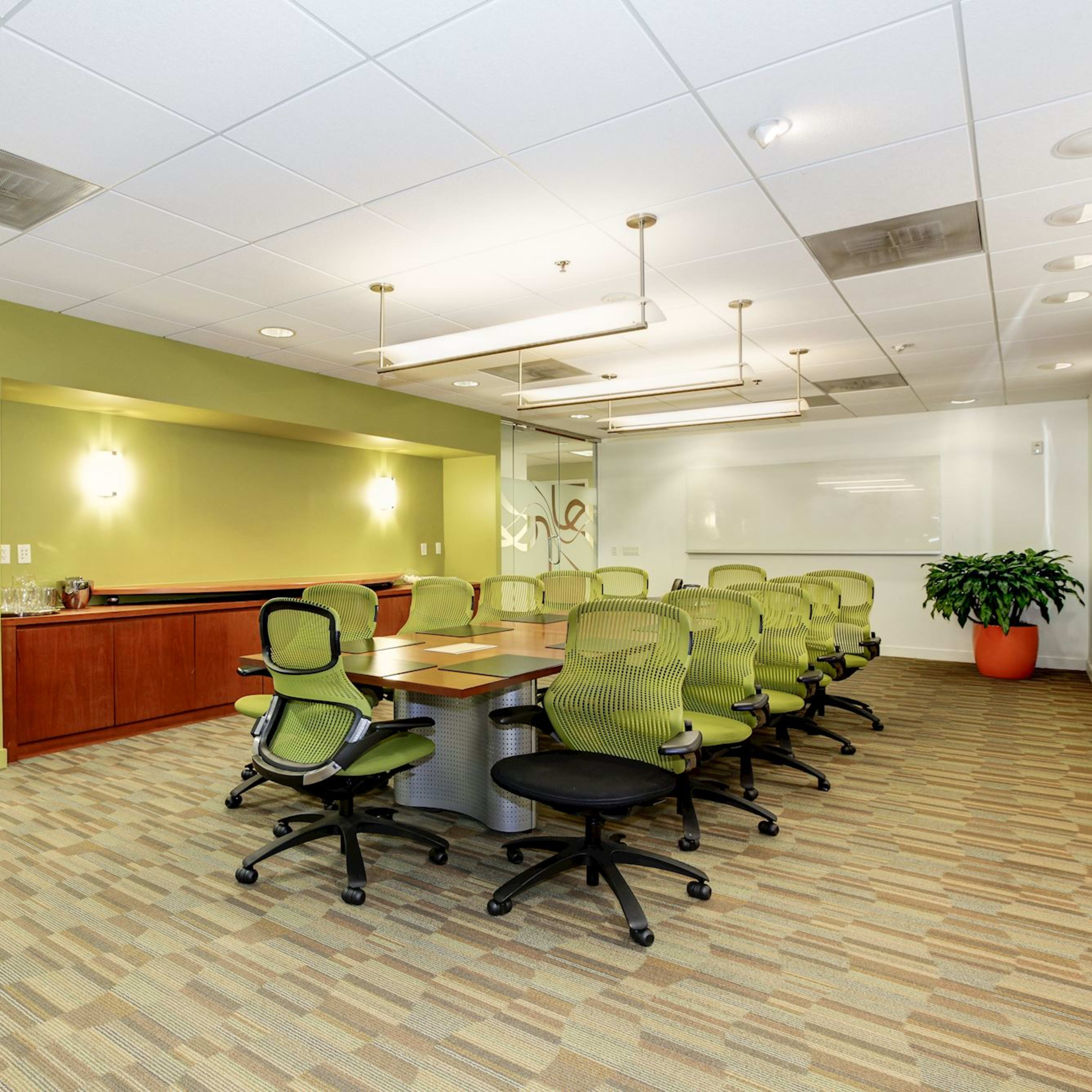 A modern conference room features a long table surrounded by ergonomic green chairs, with glass walls and a decorative plant in the corner.