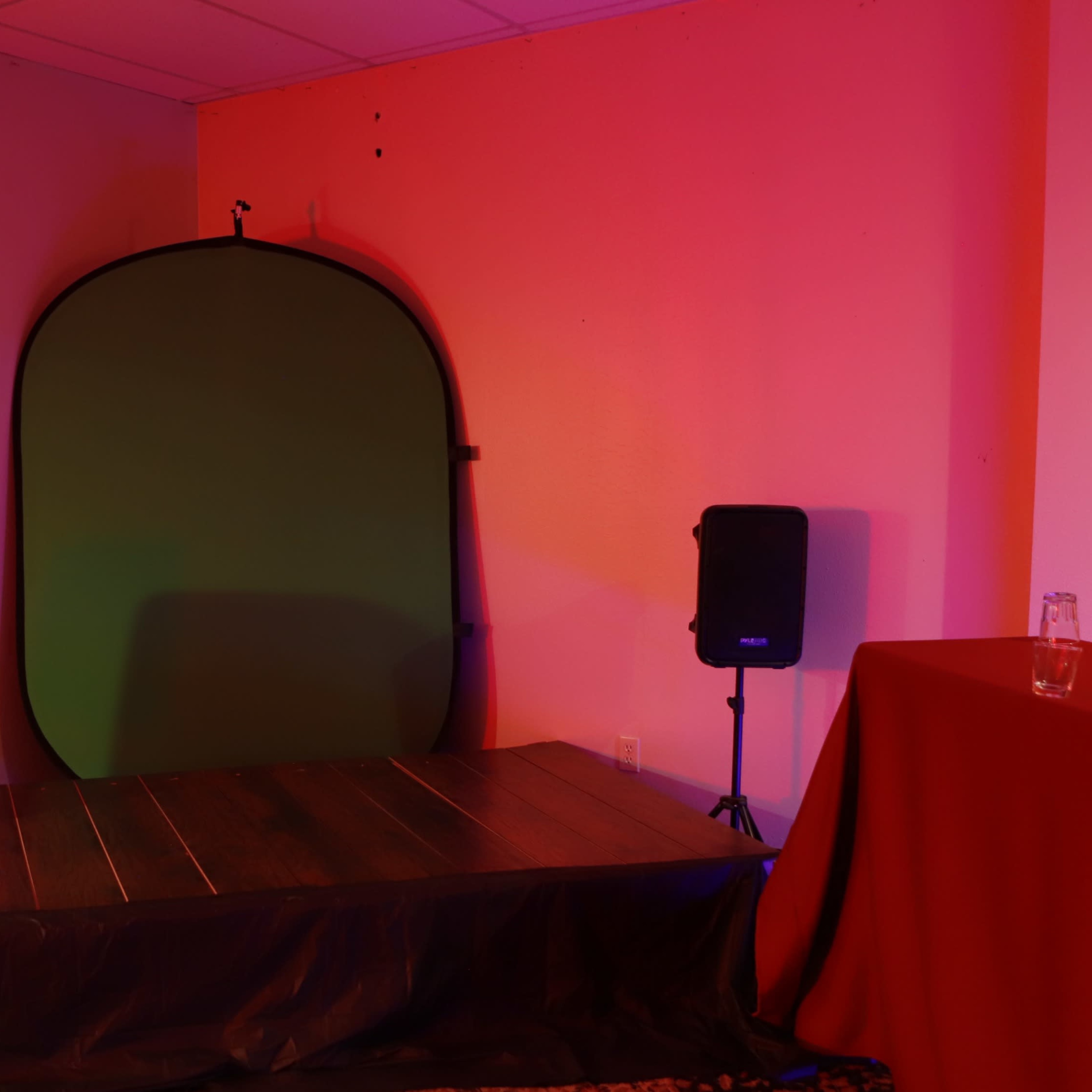 A small stage area with a green backdrop, two speakers on either side, and a table in the corner, all illuminated by colorful lighting.