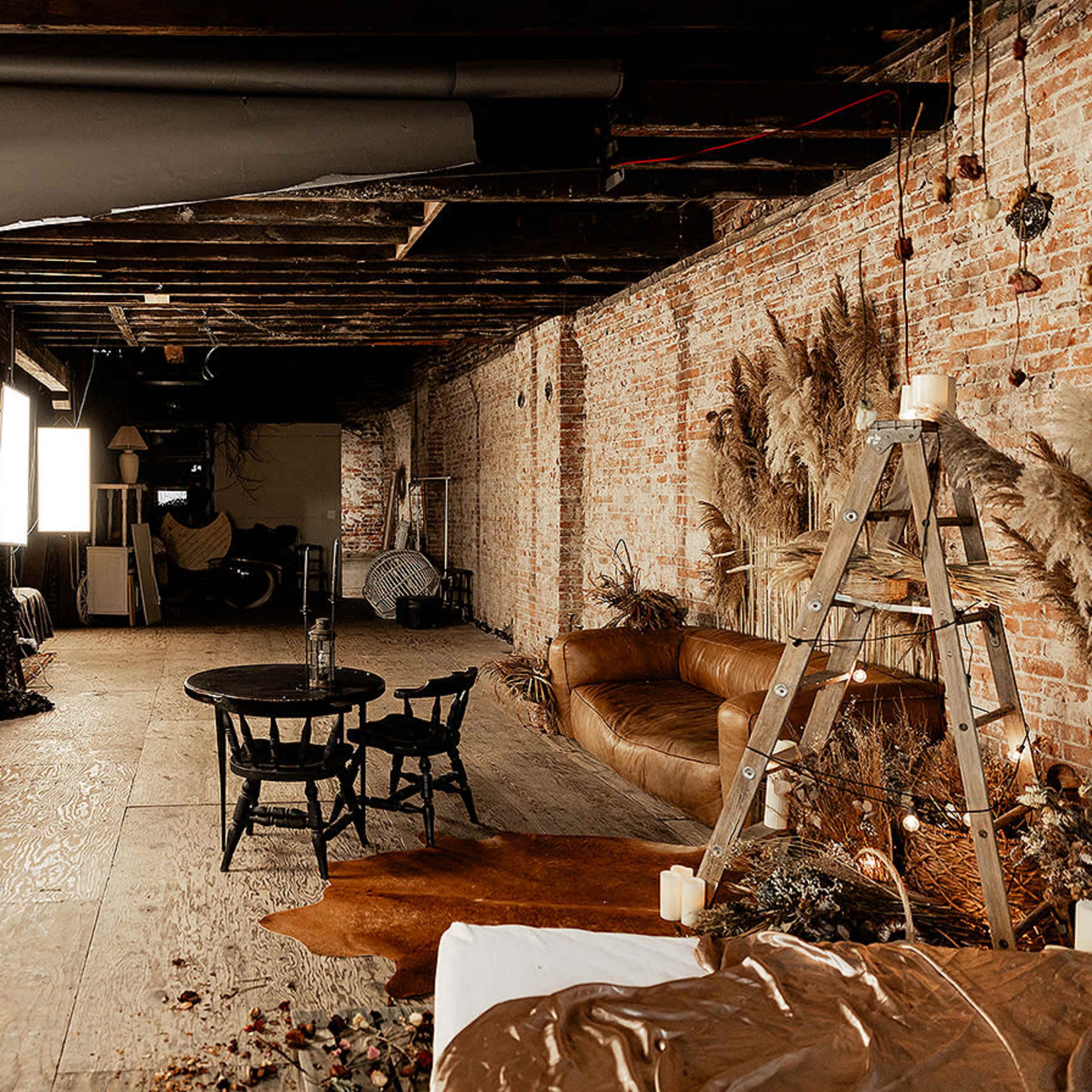 The scene shows a spacious, rustic studio with exposed brick walls, wooden floors, a brown leather sofa, a small wooden table with chairs, and various light sources set up.