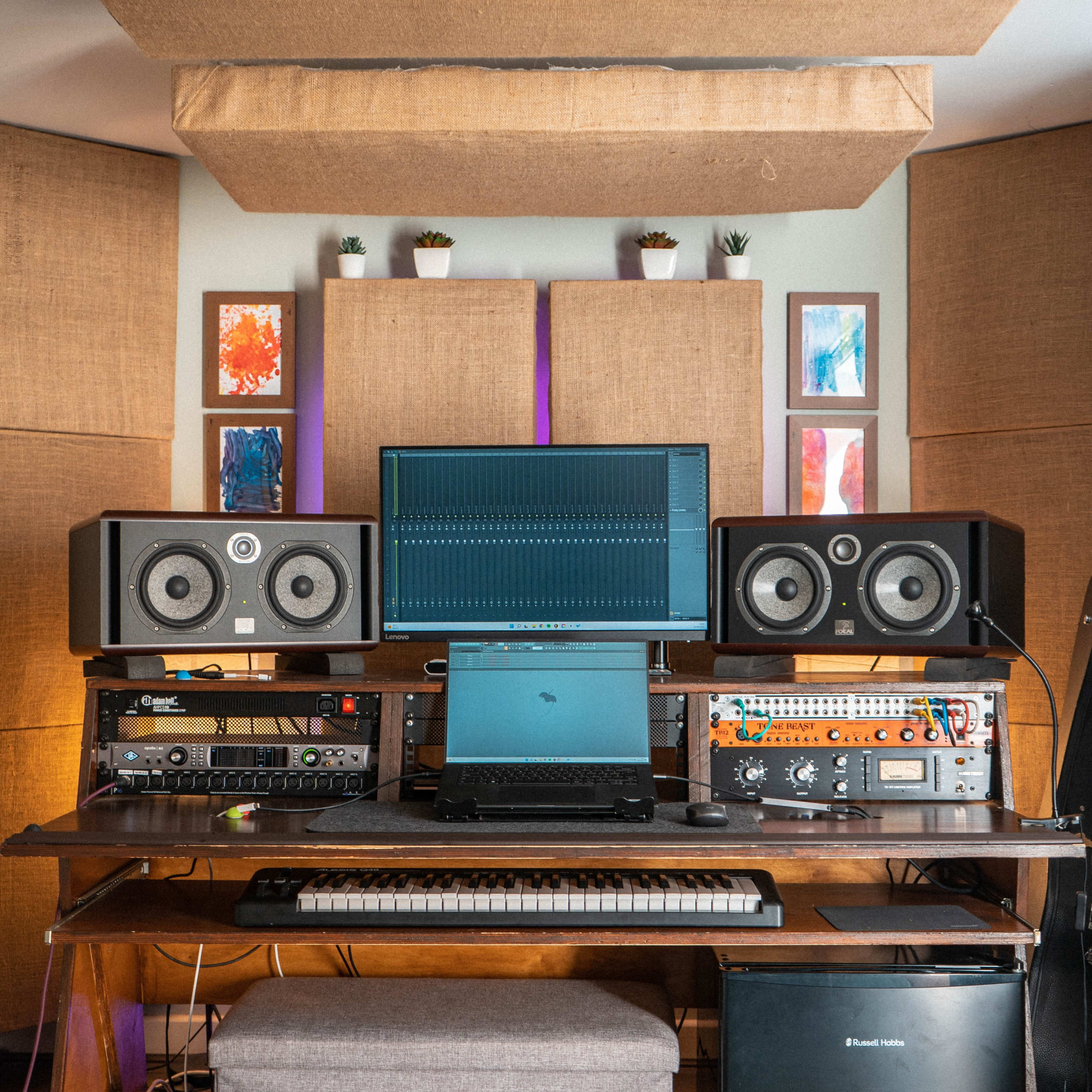 A music production workspace features a computer setup with studio monitors, an audio interface, and various other equipment arranged on a wooden desk.